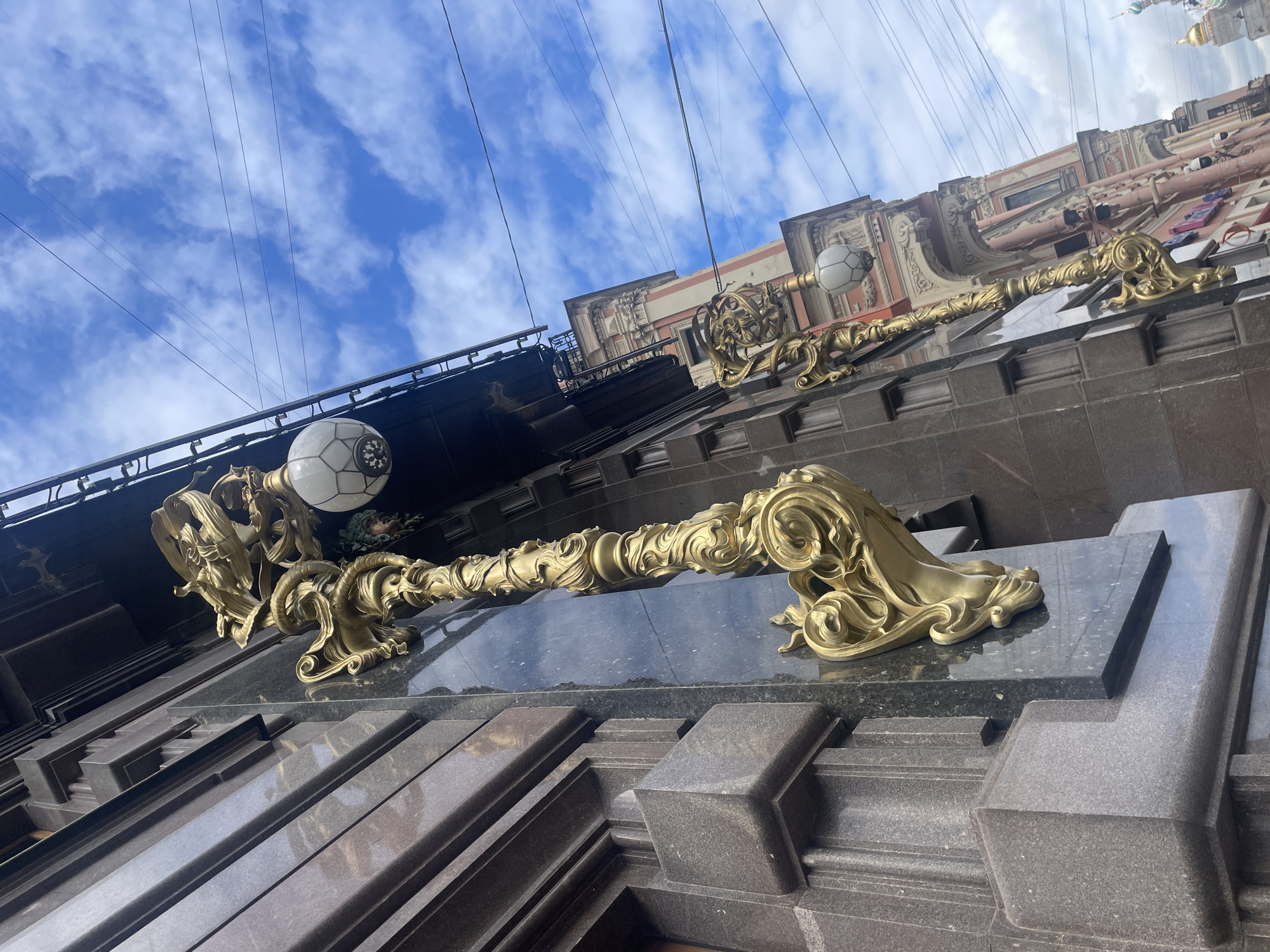 An ornate golden lamppost is mounted on the granite facade of a building in Saint Petersburg, Russia. The lamppost features intricate swirling designs and a white glass globe light fixture. Behind it, another similar lamppost is visible on an adjacent building adorned with neoclassical architectural elements in shades of pink and white. The sky above is a bright blue with scattered white clouds, and power lines crisscross the scene. The image captures a detail of the historic architecture of the city.