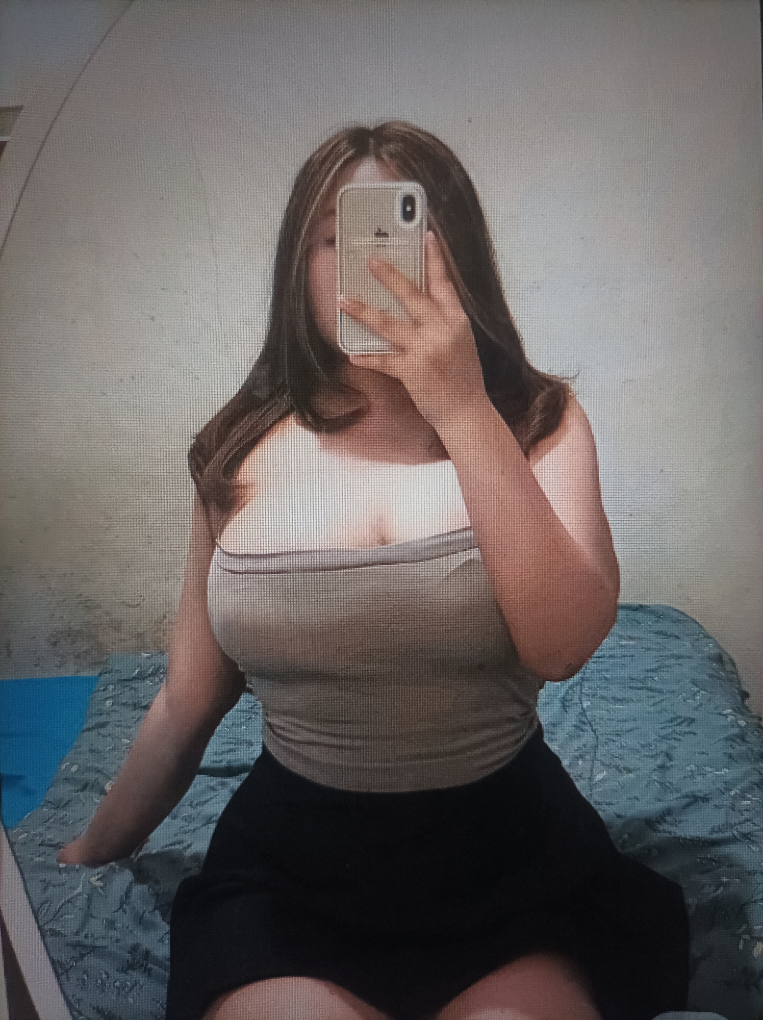 The media file shows a young person taking a mirror selfie. They are wearing a light-colored strapless top and a dark skirt. The person's face is obscured by a phone, which they are holding up to take the picture. Their hair is long and appears to be brown with some lighter streaks. They are sitting on a bed with a patterned blue bedspread. The background appears to be a wall. The lighting suggests it might be daytime, but no specific time can be determined. The overall mood is casual and personal. There is no visible text in the image.