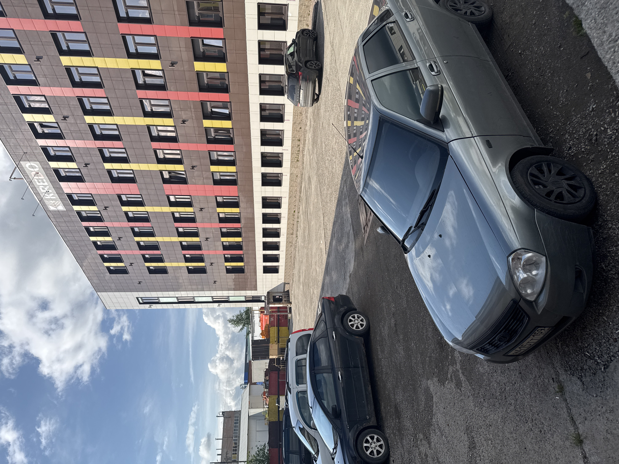 The image captures an exterior scene in Perm, Russia, featuring a multi-story commercial building, a parking lot, and industrial structures under a partially cloudy blue sky.
The primary subject is a modern, six-story building identified by the "КРЕДО" (KREDO) sign visible on its upper dark-paneled facade. This facade is predominantly dark brown, vertically accented by alternating red and yellow panels that extend the height of the building. Numerous rectangular windows with dark frames are systematically arranged across the facade. The building's base features a lighter-colored, possibly beige or white, wall section also with dark, evenly spaced windows.
In the foreground and mid-ground, a parking area with a mixed asphalt and gravel surface contains several vehicles. A grey Lada Priora sedan is positioned centrally in the foreground, displaying Russian license plate H415TT 159 (indicating Perm Krai). To the left, a black Ford Focus II sedan, a white Renault Kangoo I van, and a partially obscured dark vehicle are parked. On the right side of the parking lot, a dark grey Skoda Kodiaq SUV is parked, angled away from the viewer.
The background on the left side of the image reveals an industrial area, characterized by several low-rise buildings and structures. A prominent feature is a collection of stacked, large, open-top containers or skips in red, yellow, and blue. A small, dark-roofed structure, resembling a guard booth or utility building, is visible near these containers. Sparse vegetation, including some trees and bushes, is present. The scene is illuminated by natural sunlight, casting discernible shadows. No individuals are visible.