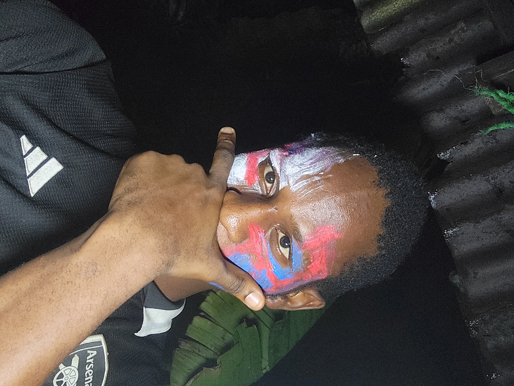 A person with face paint is photographed indoors, likely in Nkpor, Nigeria. The individual has red and blue paint around their eyes and white paint on their forehead. The person is wearing a black Adidas jacket with the Arsenal Football Club logo visible on the left sleeve. Their hand is partially covering their mouth, with one finger extended upward. Banana leaves are visible in the lower center of the image. The background appears to be a corrugated metal structure.