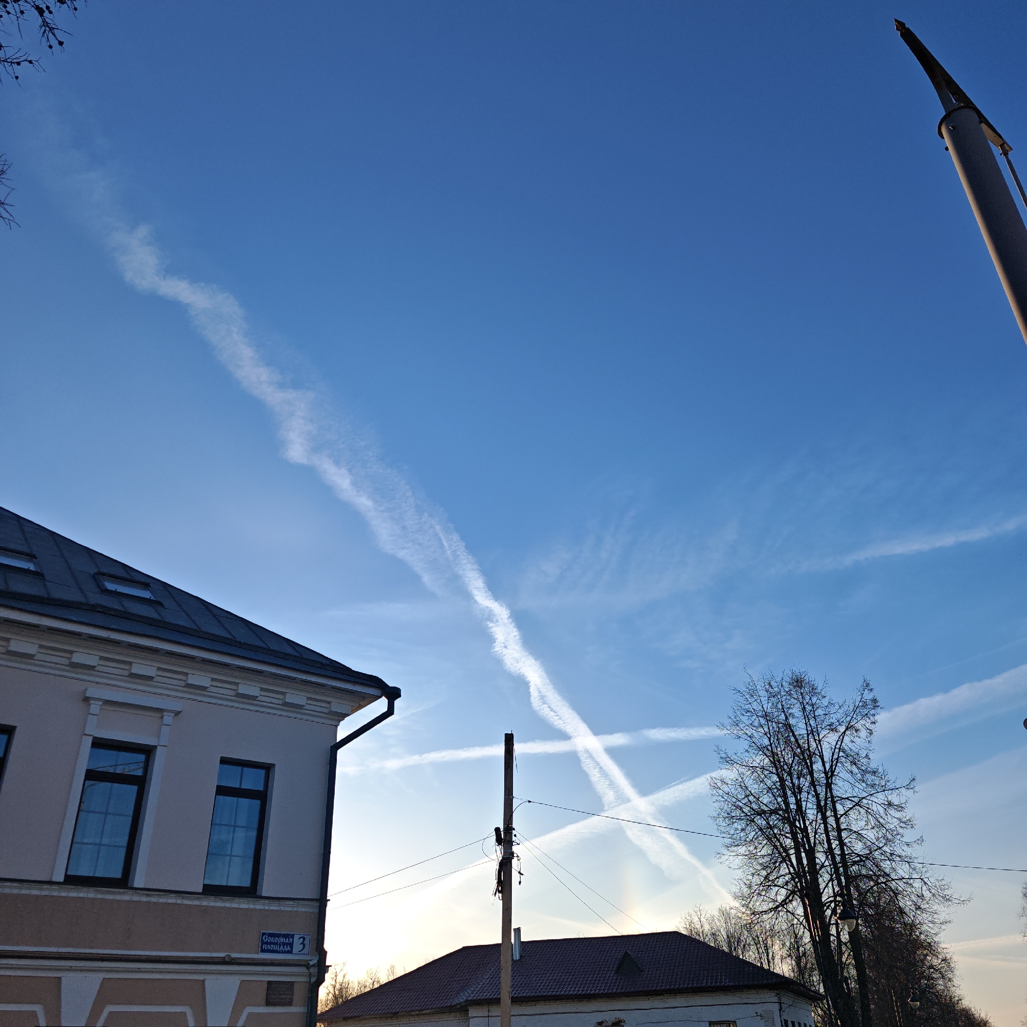 The image shows a sky with contrails forming an "X" above buildings and trees. A light pole stands between the viewer and the sky. A sign in Russian on a building reads "Соборная площадь 3" which translates to "Sobornaya Square 3," placing the scene in or near Sobornaya Square in Rostov, Russia. The sky is blue with some wispy clouds. The buildings are low-rise, with the closest one being beige and having a dark roof with attic windows. A second building is visible behind the pole with a reddish roof. There are bare trees to the right. The image is taken from a low angle, looking upward.