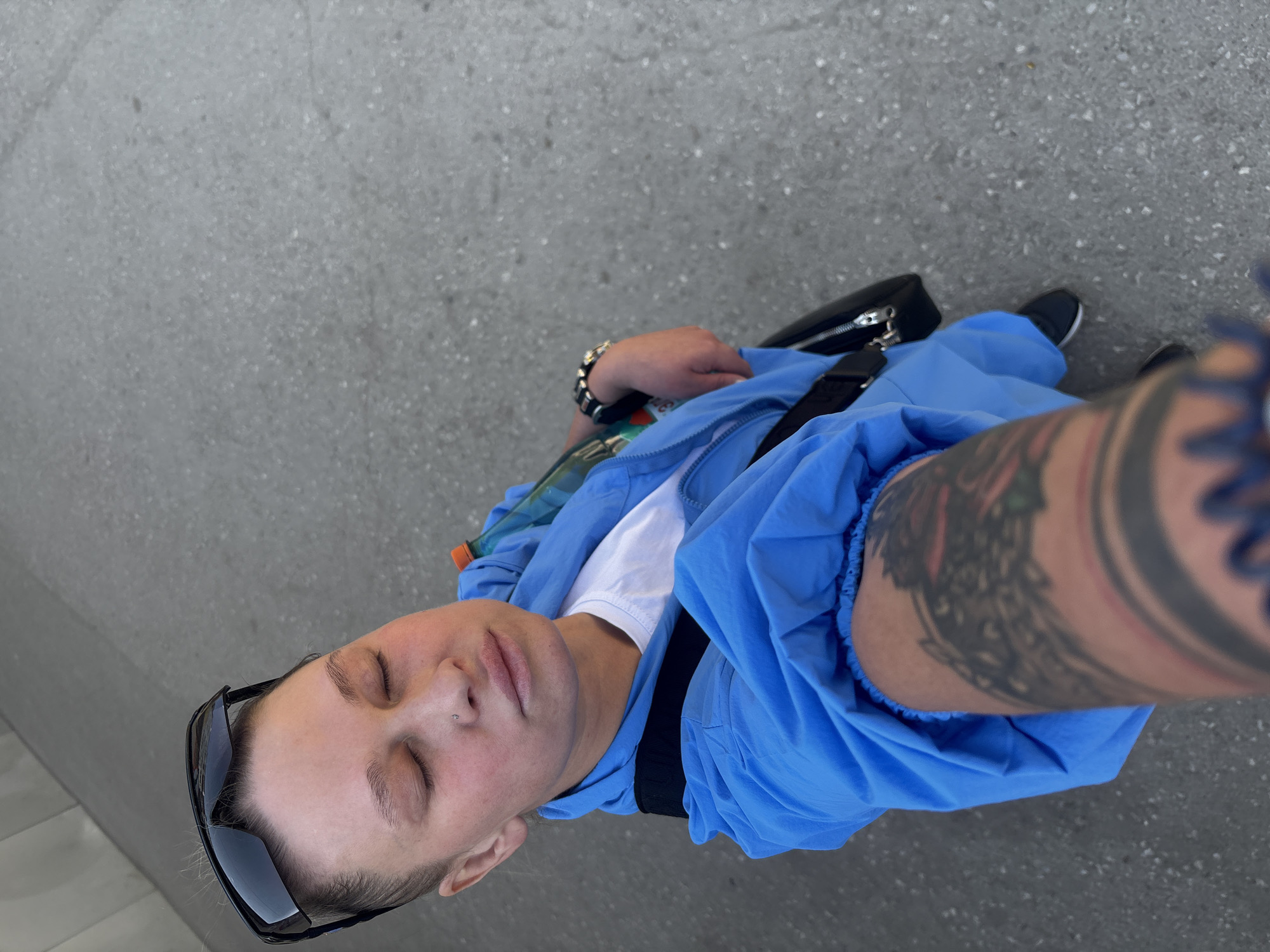 A person takes a downward-angled selfie on a gray pavement surface. The person is wearing black framed sunglasses perched on their head, a white t-shirt, and a blue zip-up jacket. A black crossbody bag with a silver zipper is slung across their body. The person has short, light hair and a nose piercing. A large tattoo is visible on the person's arm closest to the camera. Black and white athletic shoes are visible at the bottom of the frame. The location is assumed to be Perm, Russia.