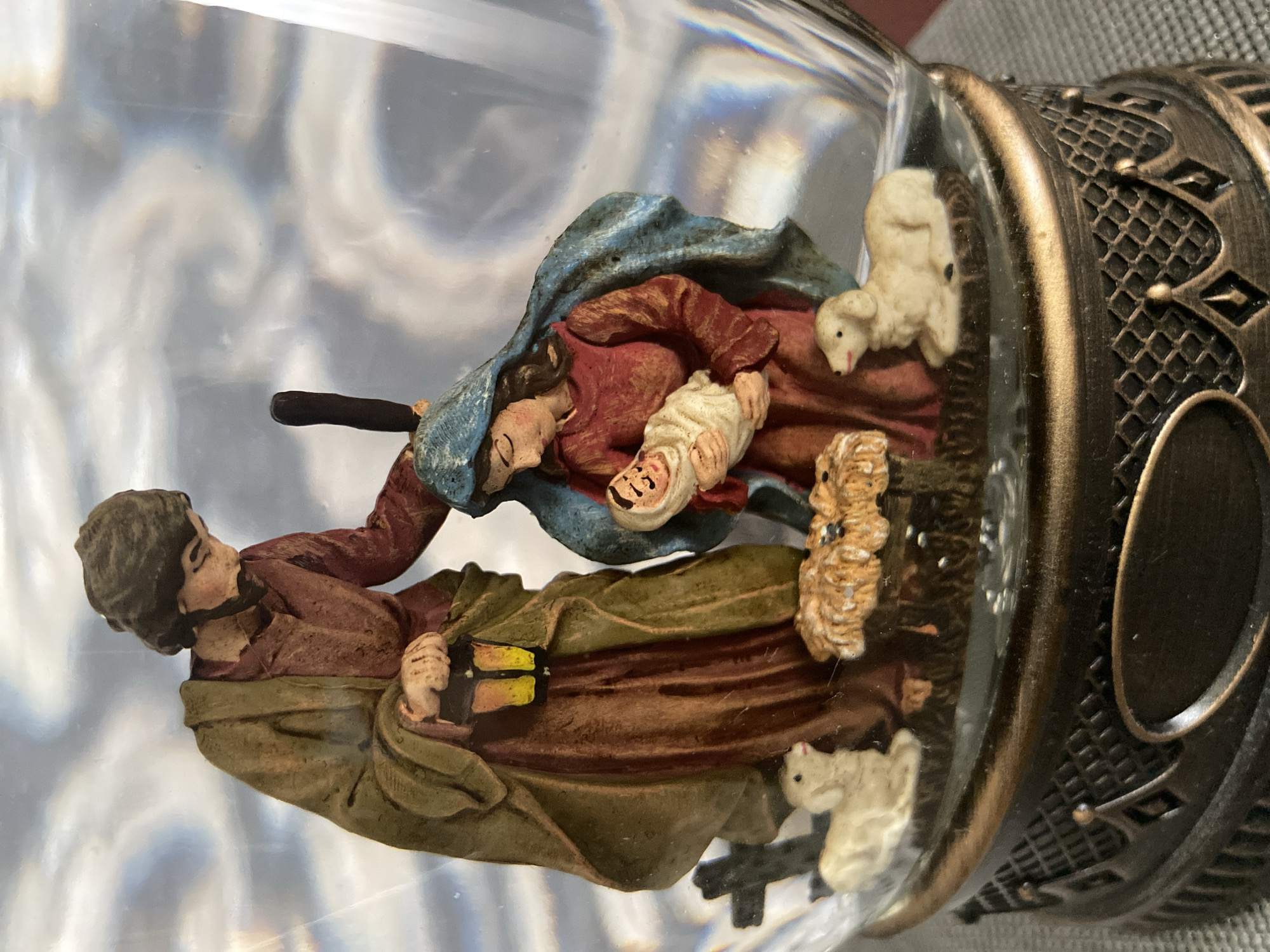 The image shows a Nativity scene figurine enclosed in a snow globe. The figurine depicts Mary holding the baby Jesus, with Joseph standing beside them holding a lantern. Two lambs are positioned at the front of the scene. The base of the snow globe is ornate and metallic. The background within the snow globe is blurry and appears to be illuminated with light and shadow. The exterior of the snow globe is transparent glass, revealing the figurine and its base. The image does not provide any information about the city of Oryol, Russia.