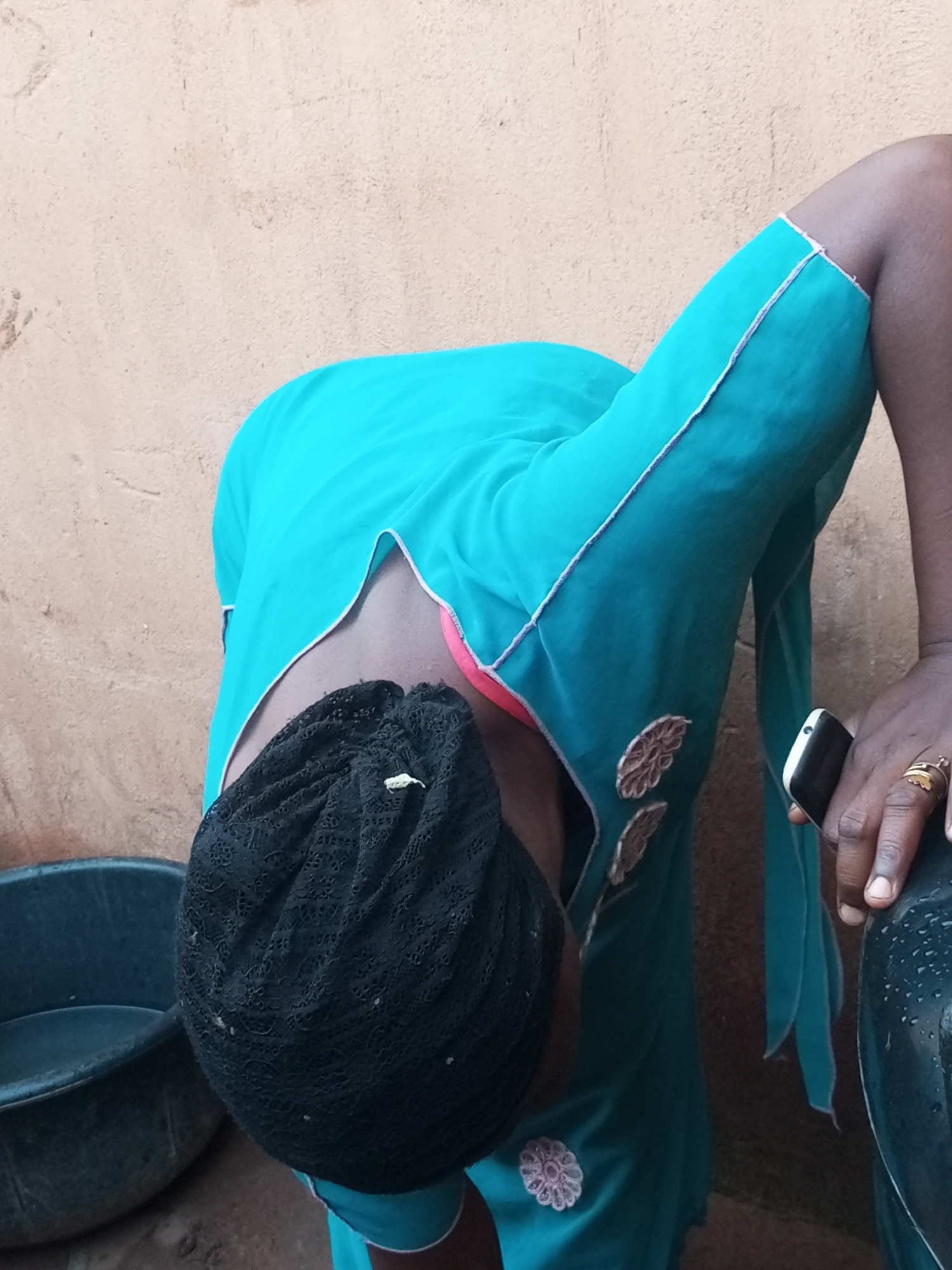 In Tempure, Nigeria, a person wearing a turquoise dress and a black lace headscarf is bent over. They are wearing a pink bra underneath the dress, and the dress has decorative floral embroidery. The person is holding a smartphone in their right hand and wearing two rings on their right ring finger. To their left is a dark gray basin. The background is a textured, light brown wall.