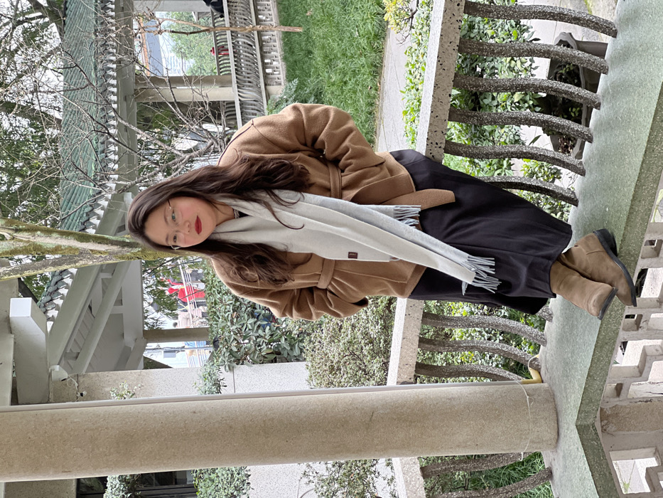 A woman with dark hair and red lipstick stands on a stone structure, dressed in a brown coat and black pants. A light gray scarf with tassels is wrapped around her neck. She is wearing tan-colored boots. The background shows greenery, trees, and a building with a traditional-style roof. The scene appears to be outdoors in a park-like setting.