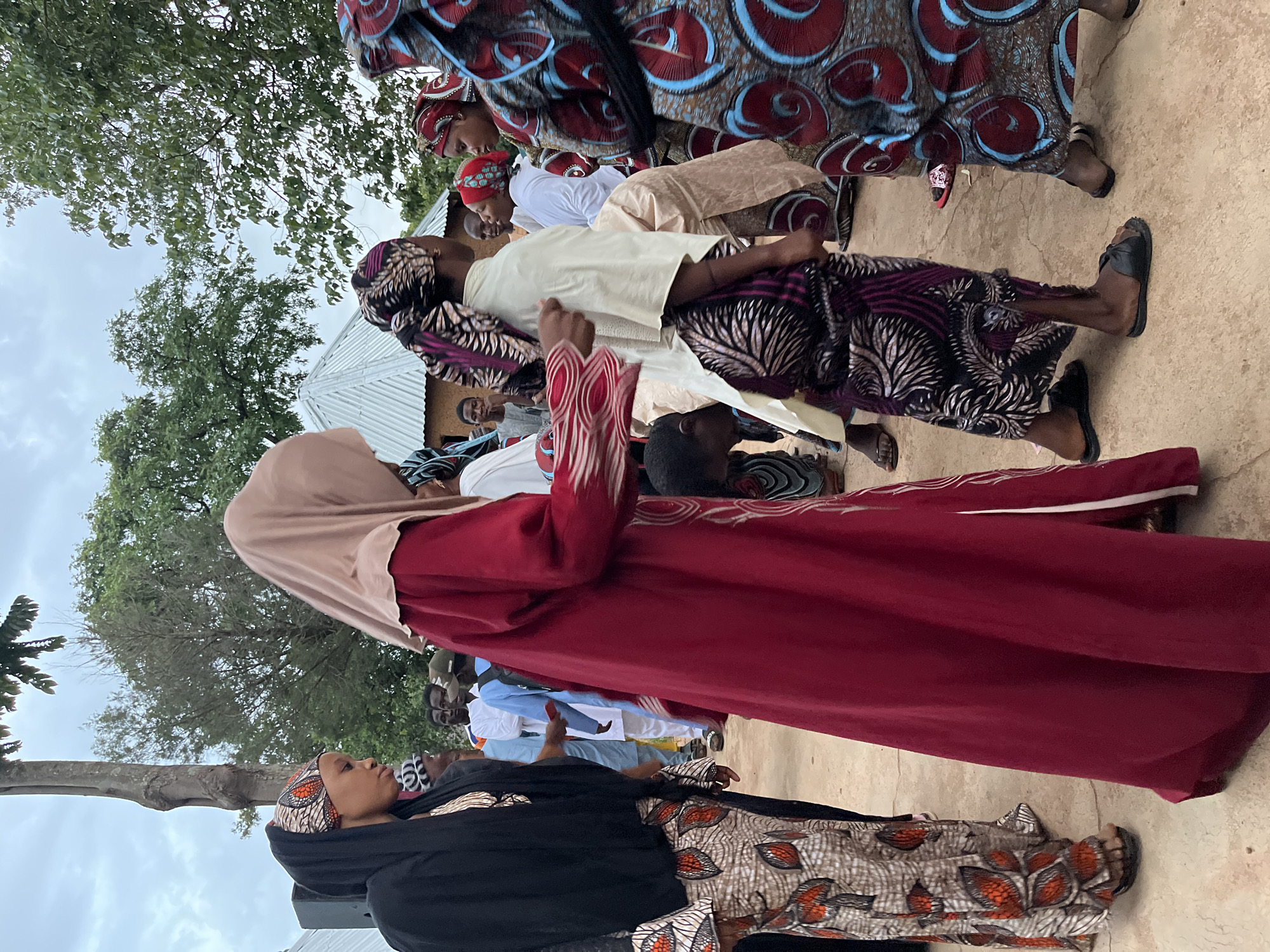 In Jos, Nigeria, a group of people are gathered outdoors. The foreground features several individuals, primarily women, adorned in colorful, patterned traditional attire, including headscarves and long dresses. One woman in the center wears a predominantly maroon dress and a light brown headscarf. To her left, another woman is dressed in black and a patterned skirt. To the right, other women are visible in vibrant, geometrically patterned outfits. Several men are also present in the background, some wearing traditional clothing and others in more casual attire. The environment appears to be a rural or semi-rural setting, with trees and simple structures, including a corrugated metal roofed building, visible in the background. The ground is a light-colored, dusty surface. The overall scene suggests a cultural gathering or event.