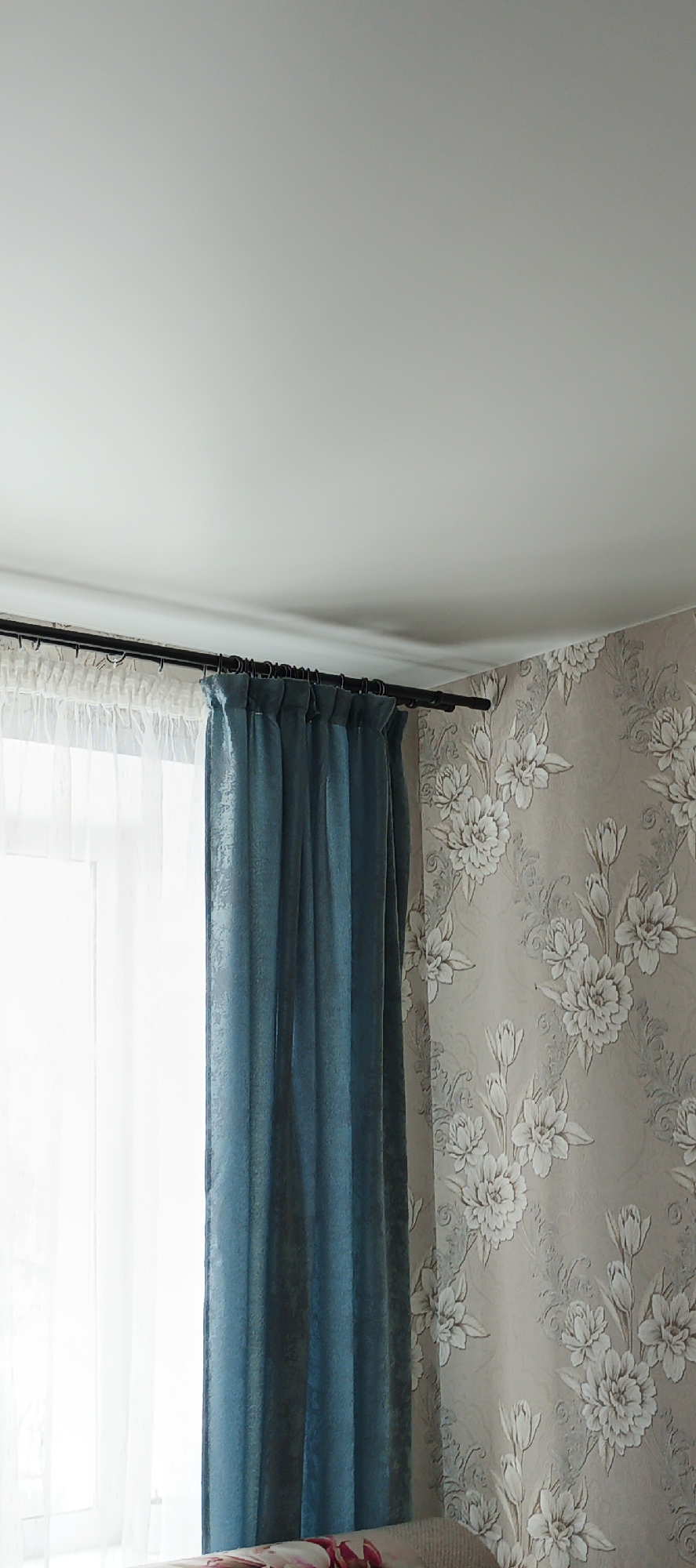This image displays an interior room located in Chaikovsky, Russia. The scene captures a section of a wall with an adjacent window and the ceiling. A window on the left side of the frame allows bright ambient light into the room and is covered by a sheer white curtain. Over this, a pleated teal-blue curtain with a subtle patterned texture hangs from a black metal curtain rod, which is secured above the window. The wall to the right is covered in light beige wallpaper featuring a repeating pattern of large, stylized white flowers and delicate grey scrollwork. The ceiling is white and flat, meeting the wall with a simple molding. The bottom edge of the image reveals a glimpse of light-colored furniture, possibly a sofa or bed, with a pink and red floral pattern. No individuals are present.