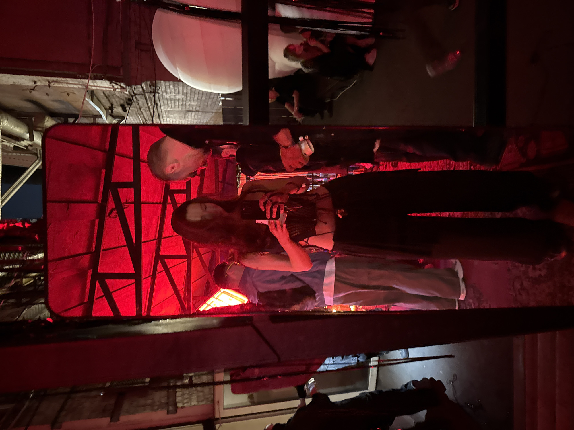 The image depicts an indoor scene in Moscow, Russia, characterized by strong red ambient lighting and an industrial aesthetic. A woman with long dark hair stands centrally, reflected in a large, dark-framed mirror. She is wearing a dark, midriff-baring top, dark wide-leg pants, and a visible chain or belt around her waist. She holds a smartphone with both hands, directing her gaze towards its screen.
Within the mirror's reflection, to the woman's right, a man with short hair and a dark top is visible, looking downwards. To the left and slightly behind the woman's reflection, another man is seen wearing a light blue jacket over a white t-shirt, light-colored pants, and glasses, with a dark backpack on his shoulders. The background of the reflection displays numerous dark structural beams and possibly scaffolding, silhouetted against the intense red illumination.
Outside the mirror's frame, on the far left, a person in dark clothing is partially visible, appearing to hold an object. On the far right, a large, rounded white object, possibly an inflatable light diffuser or decorative element, occupies the upper portion. Below it, several indistinct figures are present in the dim lighting, including one person seated on a low surface. The upper right section outside the reflection reveals exposed pipes and brickwork on a wall or ceiling, consistent with a repurposed industrial building. The floor appears dark and slightly reflective in some areas.