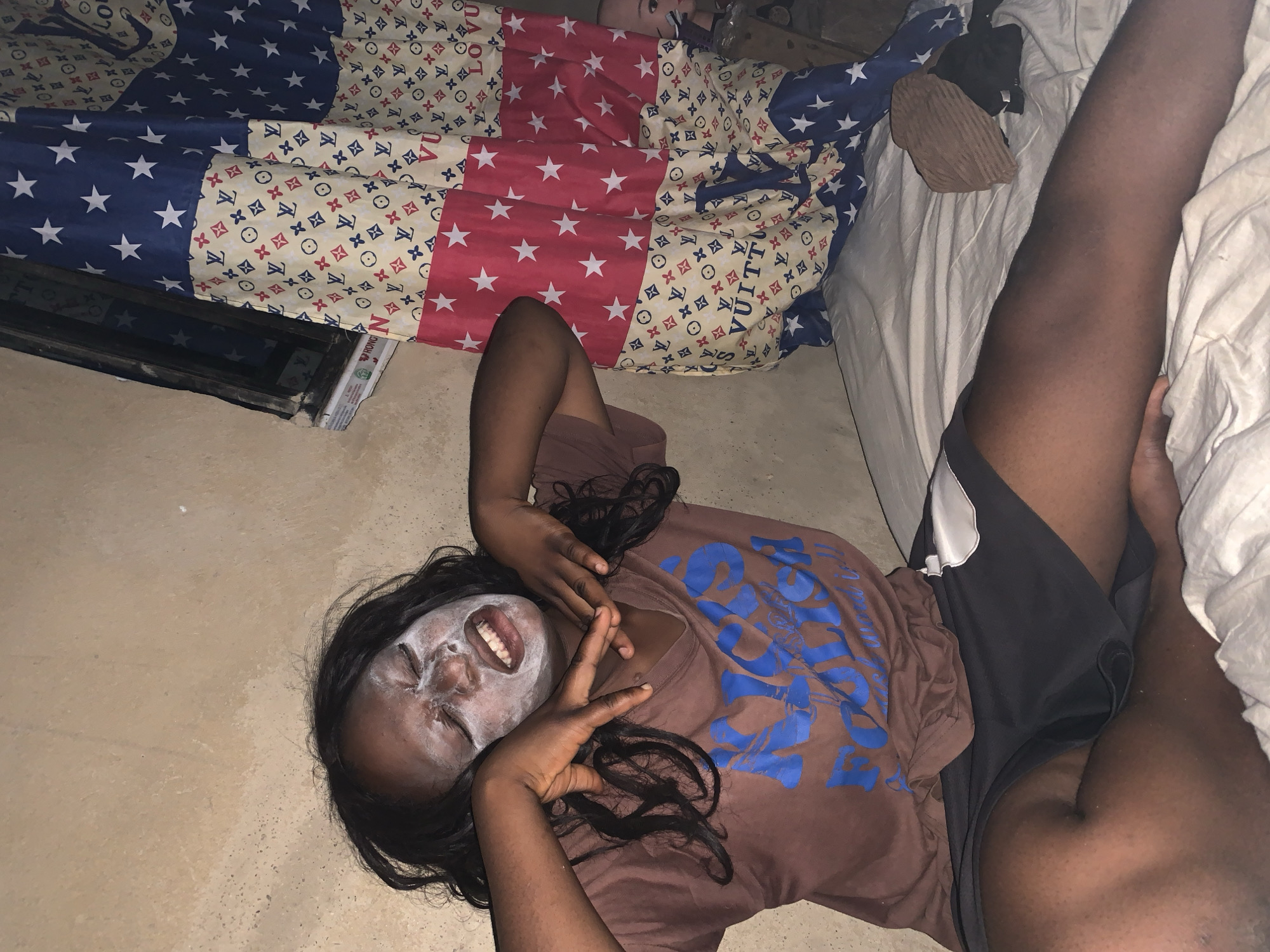 A young Black woman is lying on the floor with a white facial mask on. She wears a brown t-shirt with blue lettering and black shorts. She is making a peace sign with her hand. A bed is visible behind her, covered in a patterned comforter. The floor is beige. There is no information to identify the location in Agban, Nigeria.