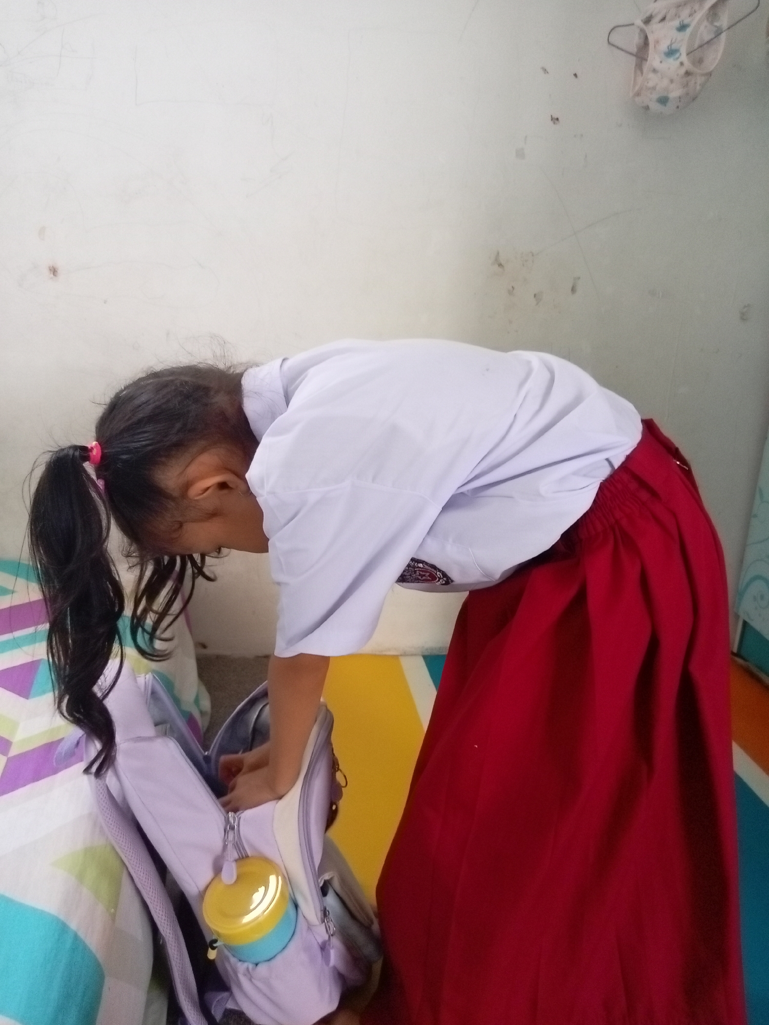 A young girl, likely of Indonesian ethnicity, is depicted from the side and slightly from the rear, bending over to place items into a light purple backpack. She has dark hair tied in a ponytail with a pink hair tie and is wearing a white collared shirt and a red skirt. The backpack has a yellow and blue cylindrical container attached to its exterior. The scene appears to be indoors, possibly a bedroom or classroom, with a white wall in the background. A colorful striped mat or bedsheet is visible on the left side of the image. The overall lighting is moderate, and there are no specific architectural features or visible signage to pinpoint a precise location within Batam, Indonesia.