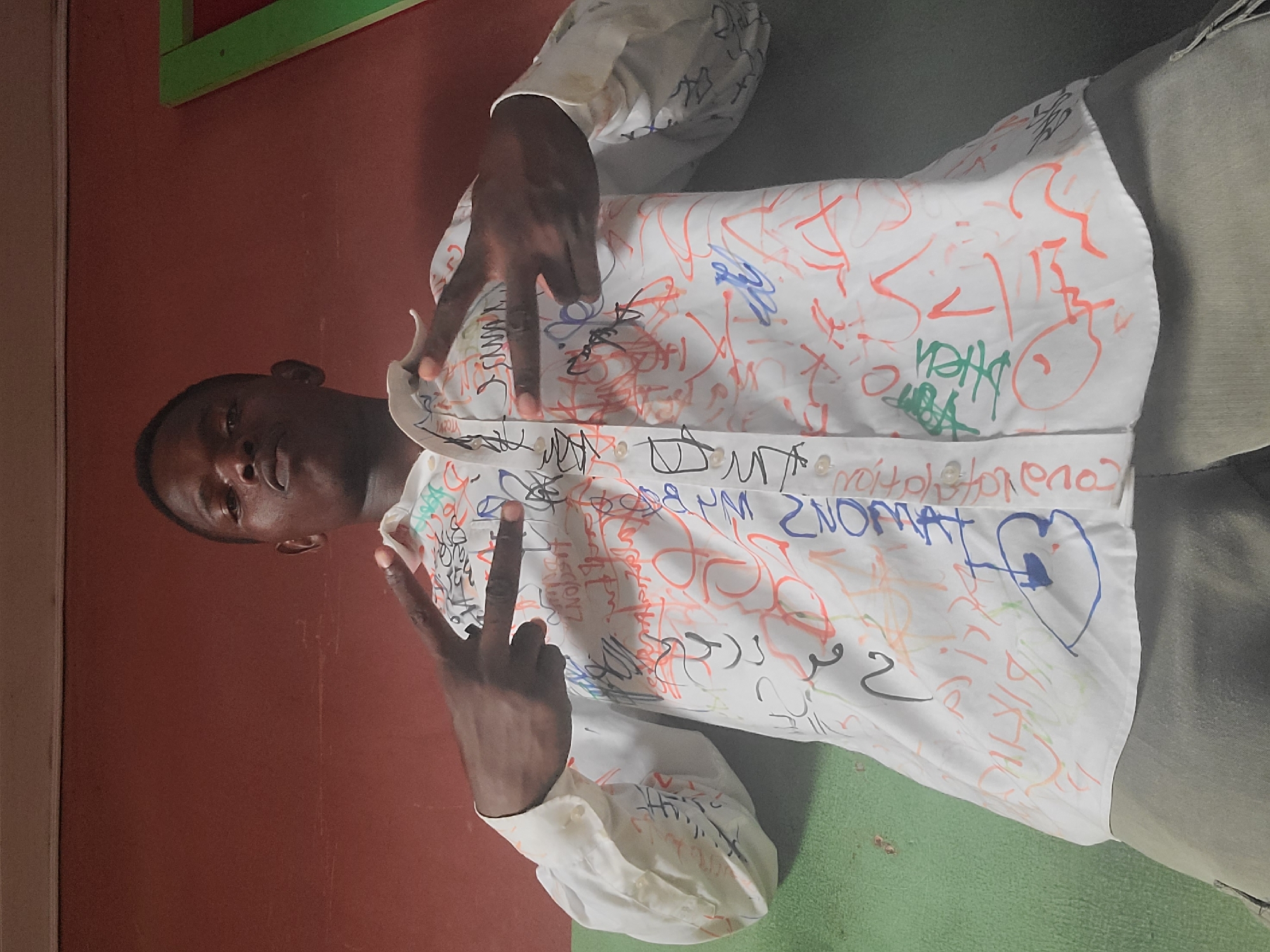 A young Black male stands in front of a reddish-brown wall and a green floor. He is wearing a white button-down shirt adorned with colorful graffiti. He holds up his hands in a peace sign. The image appears to be taken indoors, possibly in a classroom or communal area in Gura Top, Nigeria. The lighting suggests daytime. The graffiti on the shirt includes a variety of scribbled text and drawings, including a heart. The overall mood seems casual and celebratory.