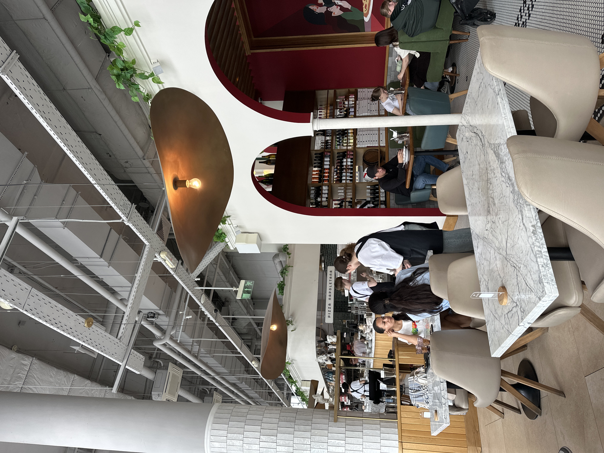 This indoor photograph captures a modern and industrial-chic restaurant or cafe interior. The high ceiling features exposed white pipes, ventilation ducts, and perforated cable trays, adorned with artificial green vines. Large, contemporary metallic pendant lights with visible bulbs hang from above, casting a warm, inviting glow.
In the foreground, two elegant, light-beige upholstered chairs flank a white marble-top table. Further into the space, several patrons are seated at various tables, some with similar marble tops and beige chairs, others with green or black seating. Activities include dining and conversation. Visible individuals include a person in a black jacket and jeans, and another in a green jacket, both seated at tables in the left midground.
Towards the right side of the image, a food preparation or serving counter is active with staff. One person in a white top and dark apron, and another in a dark top with a white apron, are seen attending to the counter. Above this area, a clear sign displays the text "PIZZA NAPOLETANA," indicating the establishment's specialty. Another individual with long dark hair is seated at a table closer to this service area.
Architecturally, the space incorporates striking deep red arched alcoves along the back wall, housing shelves stocked with numerous bottles, likely wine or spirits. One of these alcoves features a colorful mural depicting stylized human faces. The flooring varies from a light-toned surface in the foreground to a distinct black and white geometric tiled pattern under some tables further back. An emergency exit sign is also visible on the ceiling. The overall atmosphere is bustling and bright, suggesting daytime activity within a well-lit establishment.