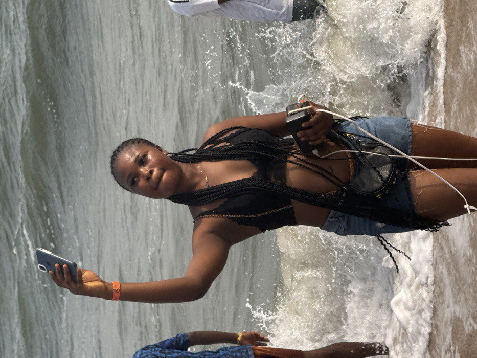 This image depicts a vibrant beach scene in Mopo Onijebu, Nigeria, with three individuals wading in the ocean. The main subject is a young woman, positioned centrally, standing in the shallow, turbulent water as waves crash around her. She has dark skin and her long black hair is styled in braids. She is dressed in a black lace bralette or crop top and light blue denim shorts, with a small black purse with a gold chain strap slung across her body.
In her left hand, the woman holds a smartphone, seemingly taking a photo or video, possibly a selfie. An orange wristband is visible on her left wrist. In her right hand, she holds another electronic device, likely a power bank, from which white cables extend, connecting to her phone or another unseen device. She has a slight smile, conveying a sense of enjoyment.
Behind her, another person is partially visible further in the water, wearing a white t-shirt and what appears to be white shorts. In the bottom foreground, a third individual is partially visible, wearing a blue patterned top and brown bottoms. The water is a greyish-green hue, with white foam and splashes from the breaking waves. The scene clearly indicates a daytime setting at a beach or coastline. There is no visible text in the image.