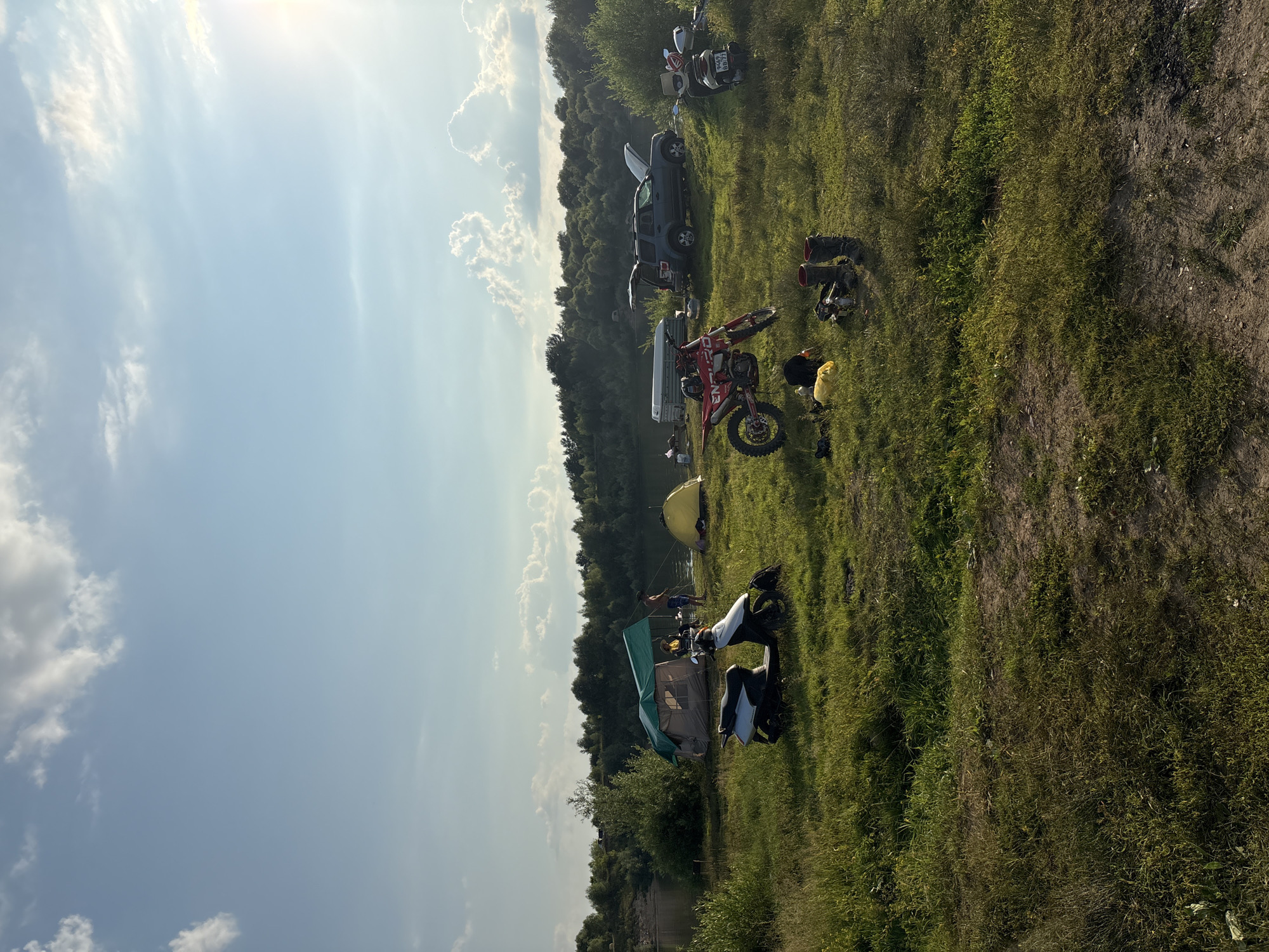 The image depicts a campsite on a grassy hillside overlooking a body of water, likely a river or lake, bordered by a dense line of trees. The sun is high in the sky, indicating it is daytime, and the weather appears to be clear with scattered clouds. The scene is set in Lan'shino, Russia.
Several objects are scattered across the landscape, suggesting an outdoor recreational gathering. There are at least two tents set up, one green and brown and another yellow. Multiple vehicles are present, including a dark-colored SUV with its doors open, a white van, and several motorcycles. One motorcycle, red and white, is prominently displayed in the foreground, leaning on its kickstand. Other objects include boots, bags, and what appears to be camping gear.
A few individuals are visible within the scene. One person is standing near the water with fishing gear. Another person is seated near a tent, and a few more are scattered around the campsite. The overall atmosphere suggests a leisurely outdoor activity, possibly camping and motorcycling. The bright sunlight and green foliage contribute to a feeling of a warm, pleasant day. There is no visible text in the image.