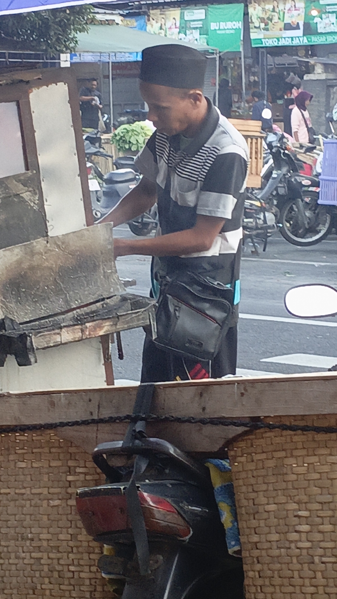 A man wearing a black kufi and a striped short-sleeved shirt with black and white horizontal stripes and grey accents stands near a food stall in Srono, Indonesia. He has a black crossbody bag with a red accent strap. In front of him is a metal grilling apparatus with visible char marks, suggesting recent use. Behind him, a street scene unfolds with motorcycles parked and people walking. A banner with Indonesian text is visible in the background. The overall environment suggests a busy market or street-side commercial area.