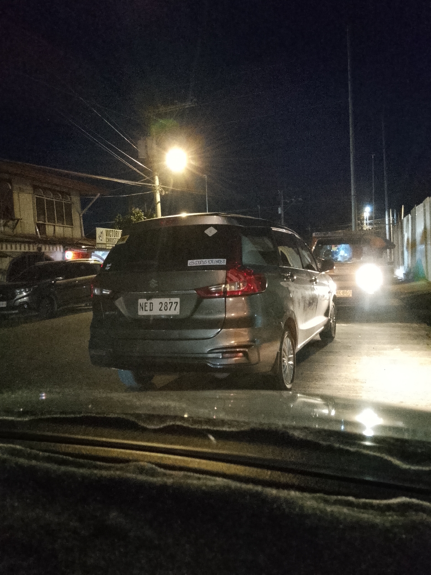 The image depicts a nighttime street scene in Davao City, Philippines. The primary subject is the rear of a grey Suzuki Ertiga, with the license plate NED 2877 clearly visible. To the left of the Ertiga, another vehicle is partially visible. In the background, a building with signs reading "VICTORY CHRISTIAN" can be seen, suggesting the location is near a church or religious institution. Streetlights illuminate the scene, and the road appears wet, reflecting the lights. The overall atmosphere is that of a typical urban street at night.