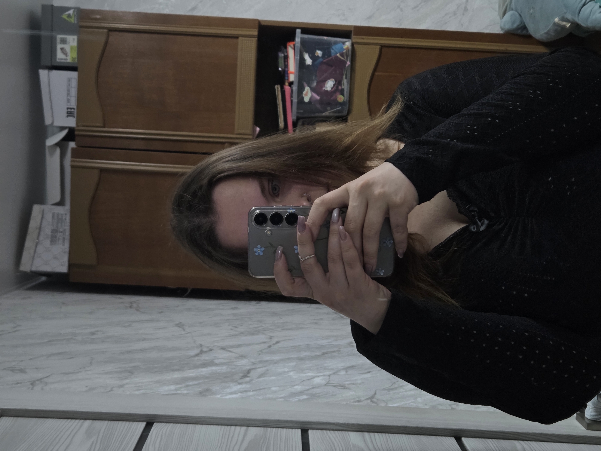 The image shows a person taking a selfie in an indoor setting, potentially a home. The person is holding a smartphone with a floral case. The background features a wooden cabinet with some items inside. The flooring appears to be marble. The person is wearing a black top. The exact location is indeterminate based on the information provided.