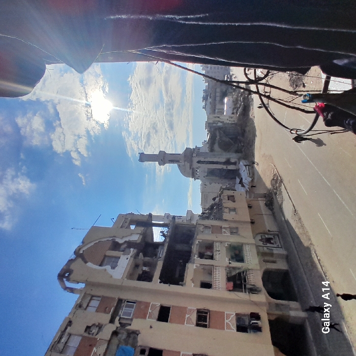 The image shows extensive destruction of buildings, including an apartment complex with significant structural damage and missing walls. A mosque with a minaret is visible amidst the wreckage. Debris litters the street. The sky is blue with scattered clouds, and the sun is shining. The scene suggests an area heavily impacted by conflict.