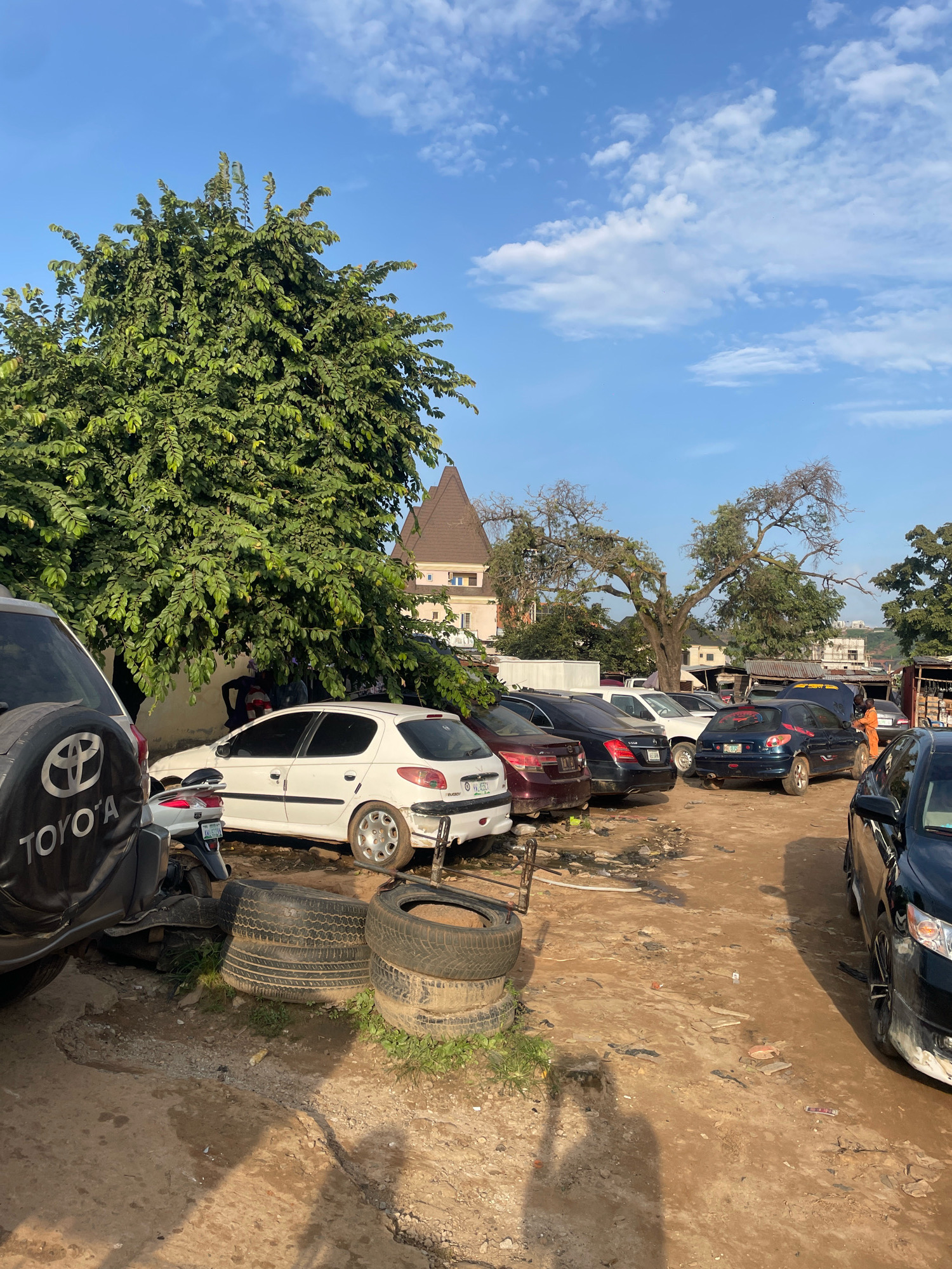The image depicts an outdoor scene in Abuja, Nigeria, possibly a car repair lot or junkyard. In the foreground, a black Toyota SUV is partially visible on the left, with its spare tire carrier displaying the Toyota logo. Next to it, a stack of used tires sits on the dirt ground. A white Peugeot hatchback is parked to the right of the SUV. Further back, several other cars are parked in rows, including a dark blue Peugeot hatchback with its hood open, and a person in an orange uniform working on it. The environment is dusty with some scattered debris. In the background, buildings and trees are visible under a bright blue sky with scattered white clouds.