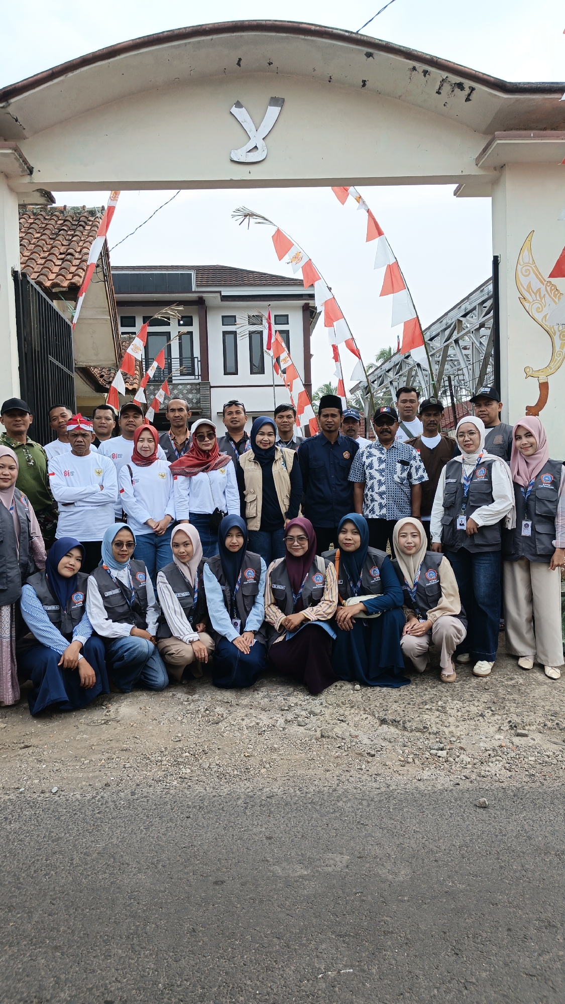 In front of a white arched entryway adorned with a stylized 'Y' emblem and flanked by red and white Indonesian flags, a group of approximately 25 people is gathered. The entryway leads to a building with a red-tiled roof and white walls. A bridge structure is visible in the background. The people are diverse in age and gender, with many wearing vests over their clothing. Some individuals are standing, while others are kneeling or crouching in the foreground. The ground in front of the group is a mix of gravel and asphalt. The scene appears to be a gathering or event in Linggasirna, Indonesia, suggested by the presence of Indonesian flags and the architectural style. The overall atmosphere suggests a sense of community or organized activity.