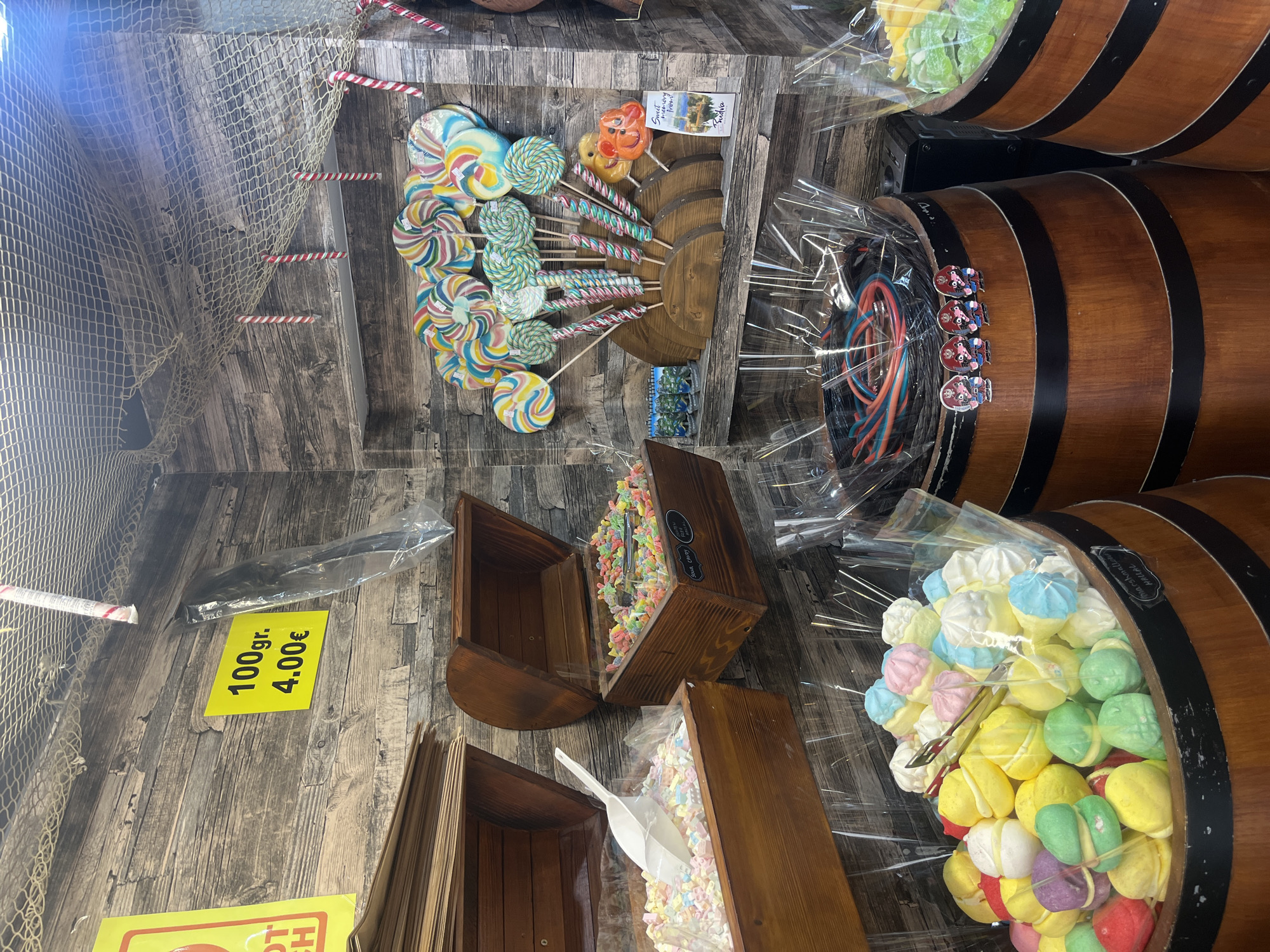 The image shows the inside of a candy shop in Budva, Montenegro. There are wooden barrels filled with assorted candies wrapped in clear plastic, including colorful marshmallows and long, multi-colored licorice ropes. Displayed on the wall are an array of lollipops with colorful swirls and spiral patterns. Below are wooden boxes filled with various candies, including alphabet-shaped marshmallows. A sign indicates a price of 4.00€ per 100 grams. A fishing net hangs on the wall for decoration. The interior is wood-paneled, giving a rustic, old-fashioned feel to the shop.
