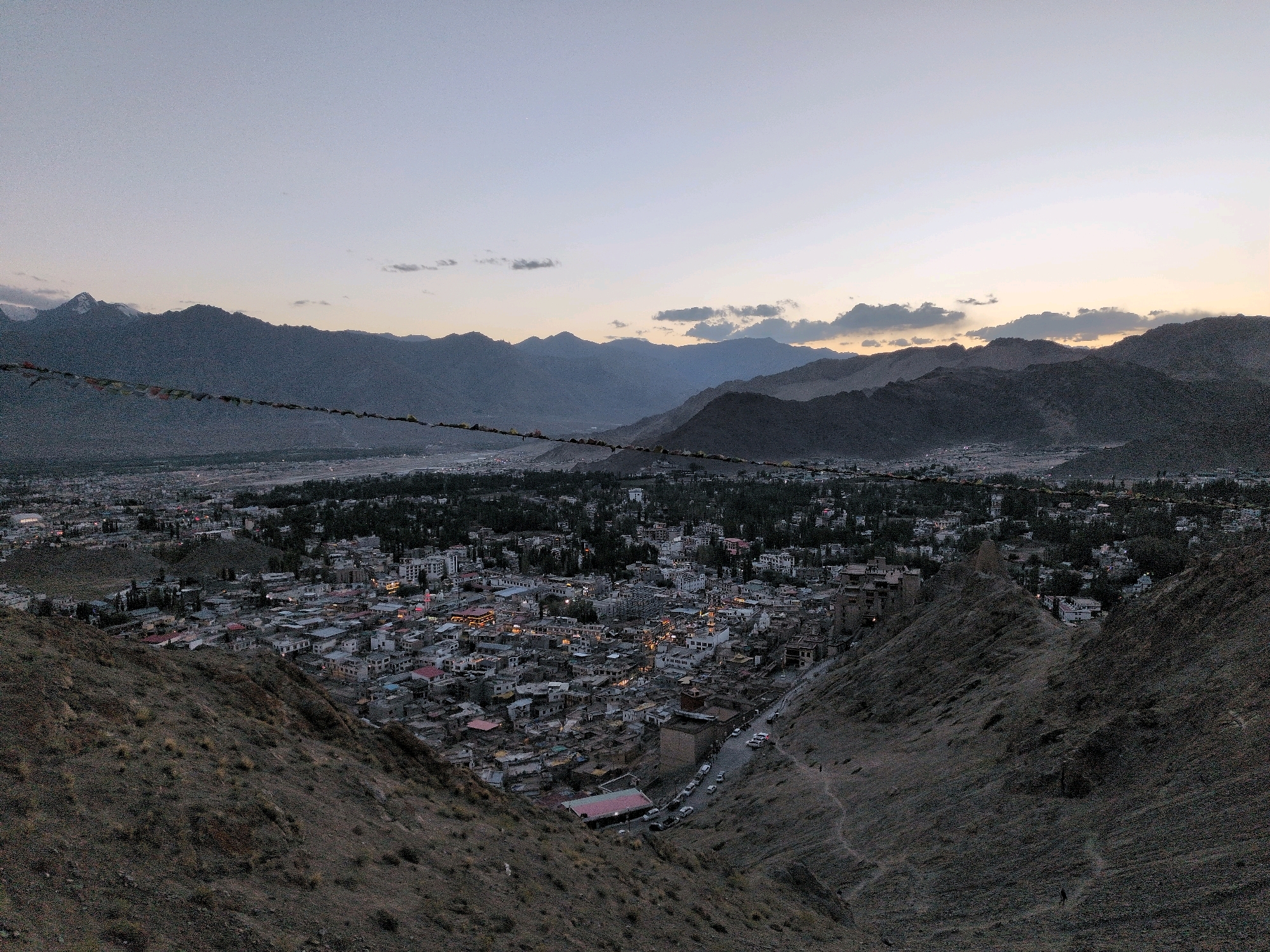 This is an aerial landscape photograph of Leh, India, taken from a high vantage point overlooking the city and the surrounding mountains. The sky is a soft gradient of blue and gray, with a hint of orange on the horizon, suggesting either dawn or dusk. Prayer flags are strung across the foreground, adding a touch of color and cultural significance to the scene.
The city of Leh is spread out below, a dense collection of buildings with predominantly white and gray facades. Many of the buildings have flat roofs and appear to be densely packed together. Lights are visible in some of the buildings, indicating that it is either evening or early morning. A road with several cars and what appear to be motorcycles winds through the city, suggesting a degree of activity. The city is nestled within a valley, with rugged, barren mountains rising steeply on either side and in the background. These mountains are characterized by their rocky, uneven terrain and lack of vegetation. The overall impression is one of a high-altitude desert city set against a dramatic mountainous backdrop.