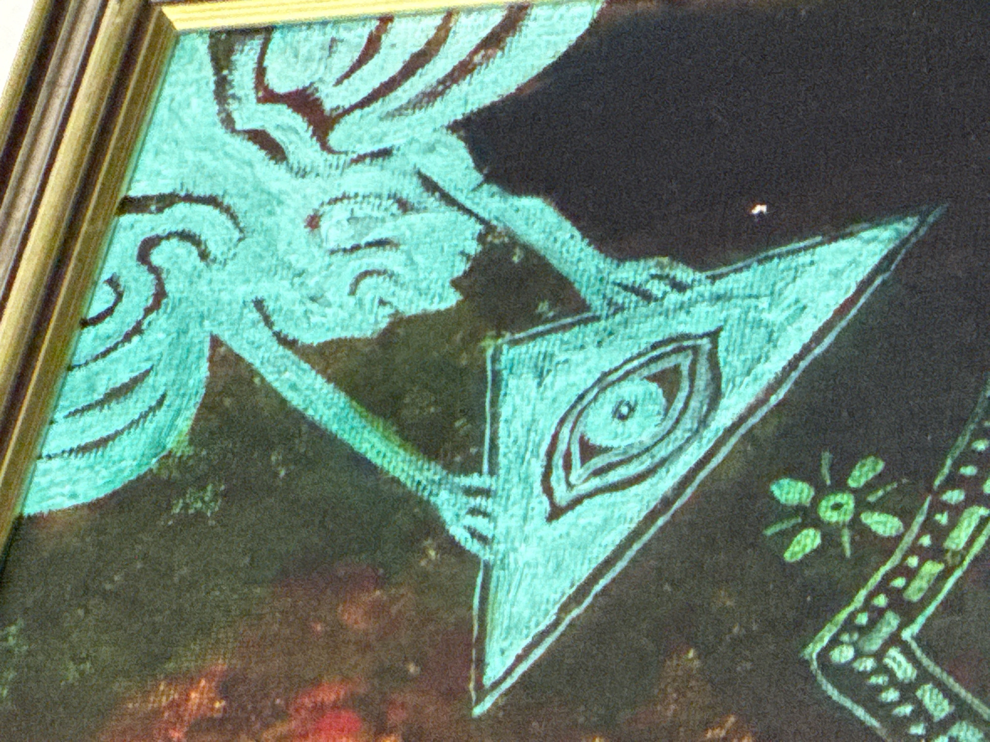 A close-up photograph shows a section of framed art with elements drawn in a teal color against a darker background. The frame itself is visible along the left edge. The artwork contains a stylized figure or creature in the upper left, holding a triangle containing an eye design. Other smaller symbols are scattered in the lower right portion of the scene, including a floral-like symbol and a design comprised of small squares in a row. The art's texture appears somewhat coarse and possibly painted. The background texture appears grainy, and a small bright spot is located in the dark background above the triangle. The exact location in the city of Plovdiv, Bulgaria is not discernible from the image.