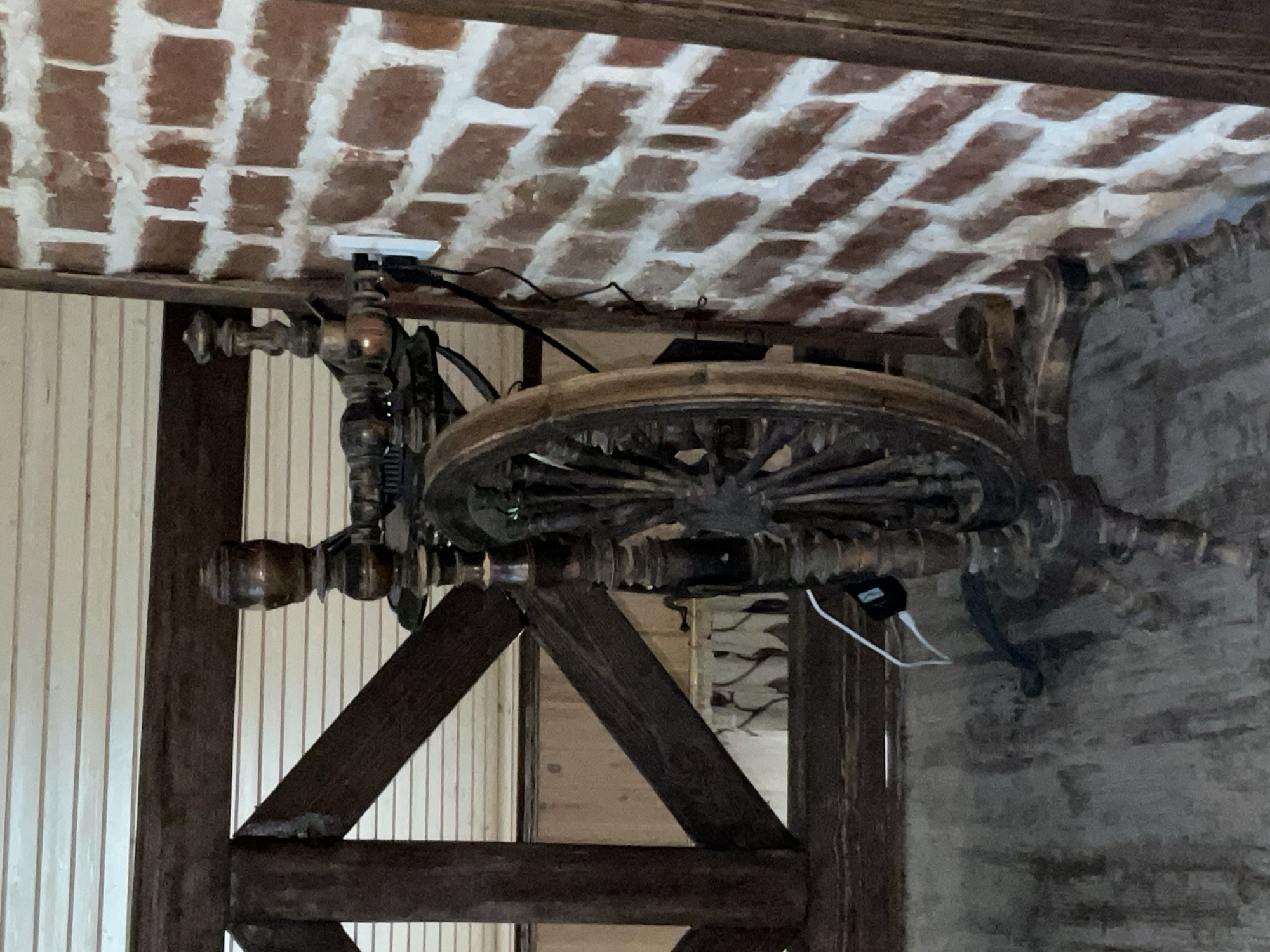This image shows an interior scene, likely a rustic or historical setting. The primary object in focus is a large, ornate spinning wheel, made of dark wood with a prominent wooden wheel. The spinning wheel appears to be part of a larger wooden structure, possibly a table or a display.
Above the spinning wheel, the ceiling is visible, constructed with a pattern of red bricks and white mortar. The bricks are laid in a staggered, repeating pattern.
To the left of the spinning wheel, a wall covered in vertical white wooden planks can be seen. This wall suggests an indoor environment with a rustic or country aesthetic.
On the spinning wheel's mechanism, several electrical devices are attached. A black power adapter with a white cord is plugged into what appears to be a power strip or outlet, which is mounted beneath the brick ceiling.
The overall impression is one of an antique or decorative item placed within a space that embraces a rustic or aged ambiance. There is no visible text in the image. The lighting appears to be artificial, and there are no indications of the time of day or weather conditions. The scene is static, with no apparent activity taking place.