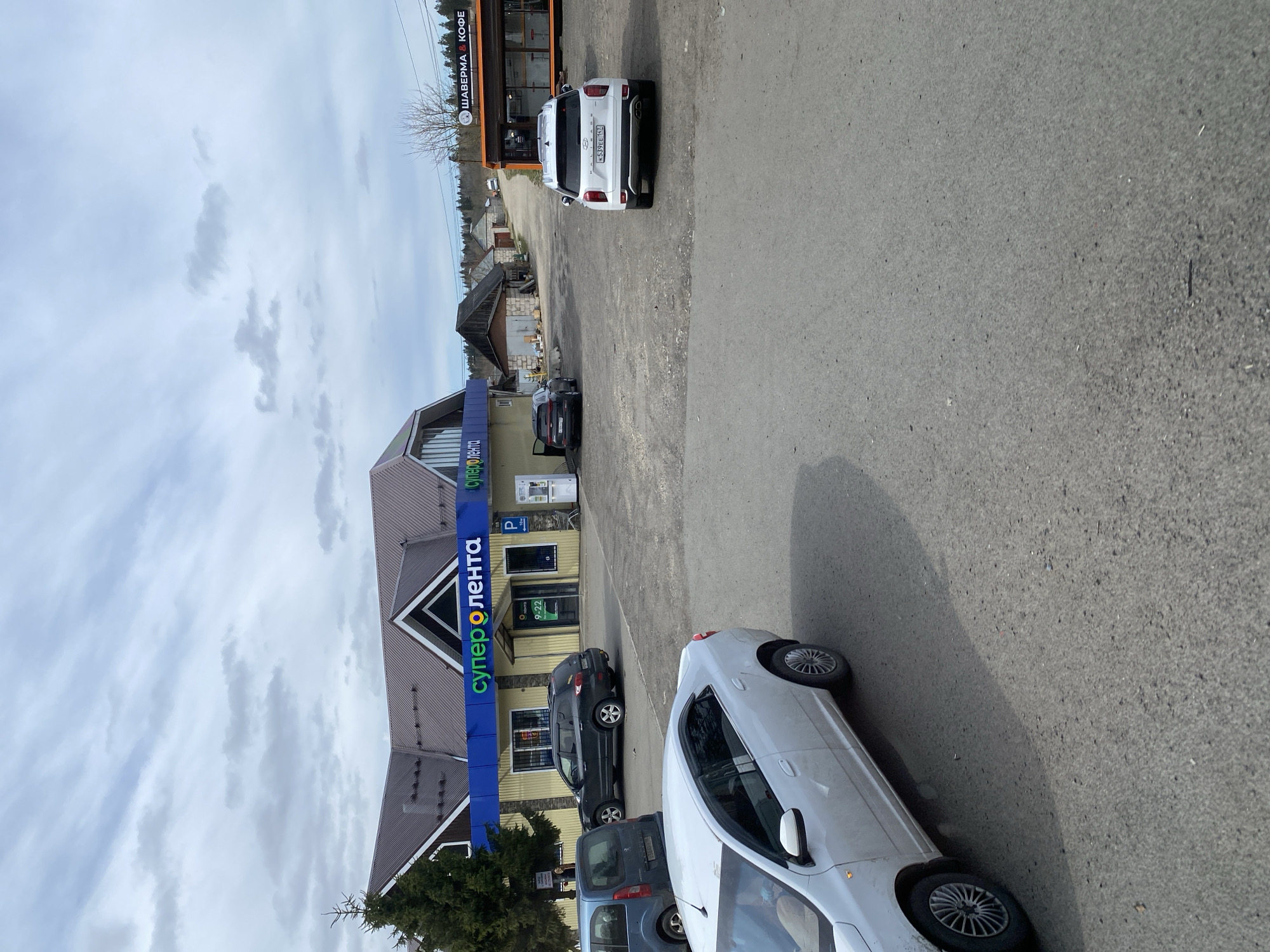 The image captures an outdoor scene at a commercial location in Seltso, Russia, under an overcast sky. In the foreground, an asphalt road occupies the right side, with a white car partially visible. To the left, a dirt parking area leads to two main buildings.
The most prominent structure is a supermarket with a blue facade and a distinctive brown, multi-gabled roof, featuring the large sign "СУПЕР O ЛЕНТА" (Super O Lenta). Parked in front of this building are a dark-colored SUV and a blue car. Further in the background, another, smaller building with an orange-brown facade houses a cafe. A white Hyundai Creta SUV, bearing the Russian license plate M539EE 174 (indicating Chelyabinsk Oblast), is parked near this cafe.
The activity depicted is typical for a commercial area, with cars parked for patrons visiting the stores or cafe. The cloudy sky suggests an overcast day, and some bare trees in the background imply it might be late autumn, winter, or early spring. A coniferous tree stands near the supermarket entrance.
Visible text includes: "СУПЕР O ЛЕНТА", a "P" parking sign, "OPEN", and "9-22" (indicating store hours) on the supermarket building. The cafe building displays "ШАВЕРМА & КОФЕ" (Shawarma & Coffee).
