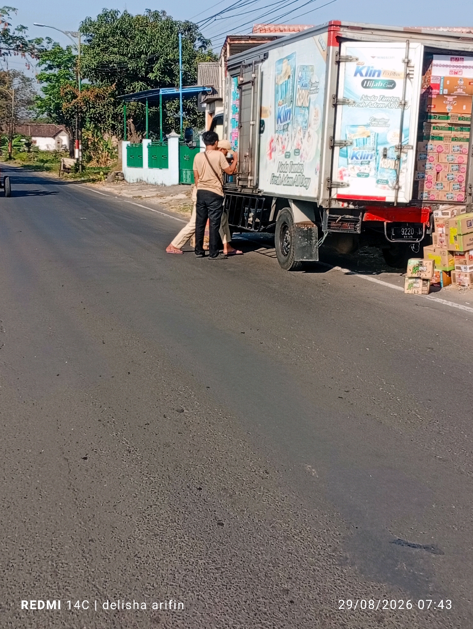 A two-person crew is seen by the side of a paved road in Karang Asem, Indonesia, near a parked delivery truck. The truck, identified by a license plate that reads "L 9220 W", is filled with stacked boxes, many of which are branded with the "Klin" logo, suggesting they contain laundry detergent or similar household products. One individual, wearing a tan shirt and black pants, is standing next to the open rear doors of the truck, while the second person, in light-colored pants and a dark top, appears to be assisting with the unloading or arranging of the goods. The scene is illuminated by bright sunlight, casting distinct shadows on the road surface. In the background, residential buildings with tiled roofs and lush green trees are visible, indicative of a semi-urban or suburban setting. The environment suggests a warm, clear day.