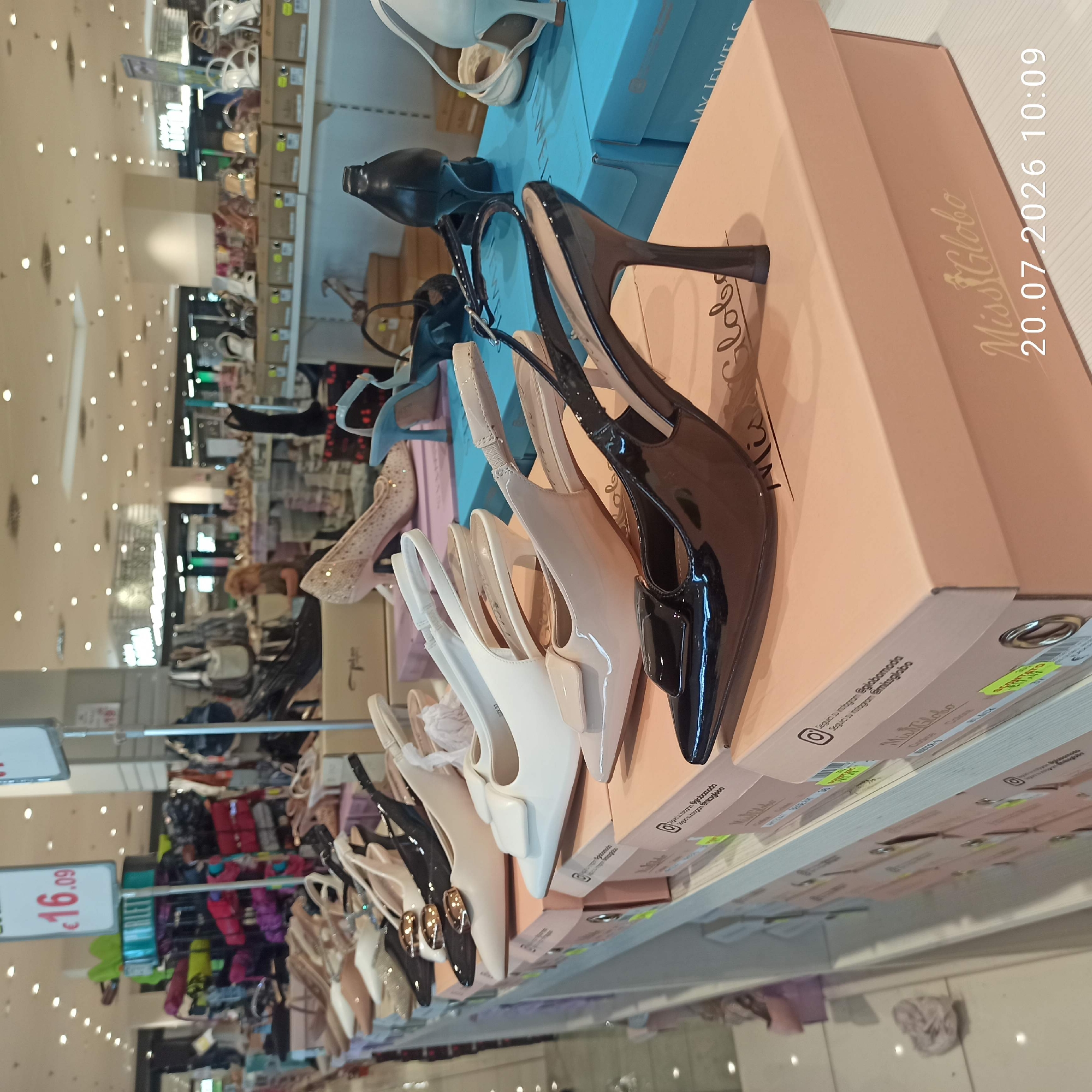 The image shows a retail store display of women's shoes in what appears to be a shopping mall. In the foreground, a row of high-heeled shoes in various colors, including black patent leather and nude patent leather, are neatly arranged on top of their respective shoe boxes. The boxes are light pink and bear the "Miss Globo" logo, along with Instagram handles. A price tag of "€16.09" is visible on a white sign in the upper left corner, suggesting a sale or promotion. In the background, more shoes and accessories are displayed on shelves and racks. Some people are visible in the mid-ground and background, browsing the merchandise, including a woman with blonde hair in the middle distance. The lighting suggests an indoor environment with overhead fluorescent lights. While the specific location in Bari, Italy is not explicitly stated, the presence of Italian currency and the context of a shopping mall are consistent with a commercial establishment in the city.