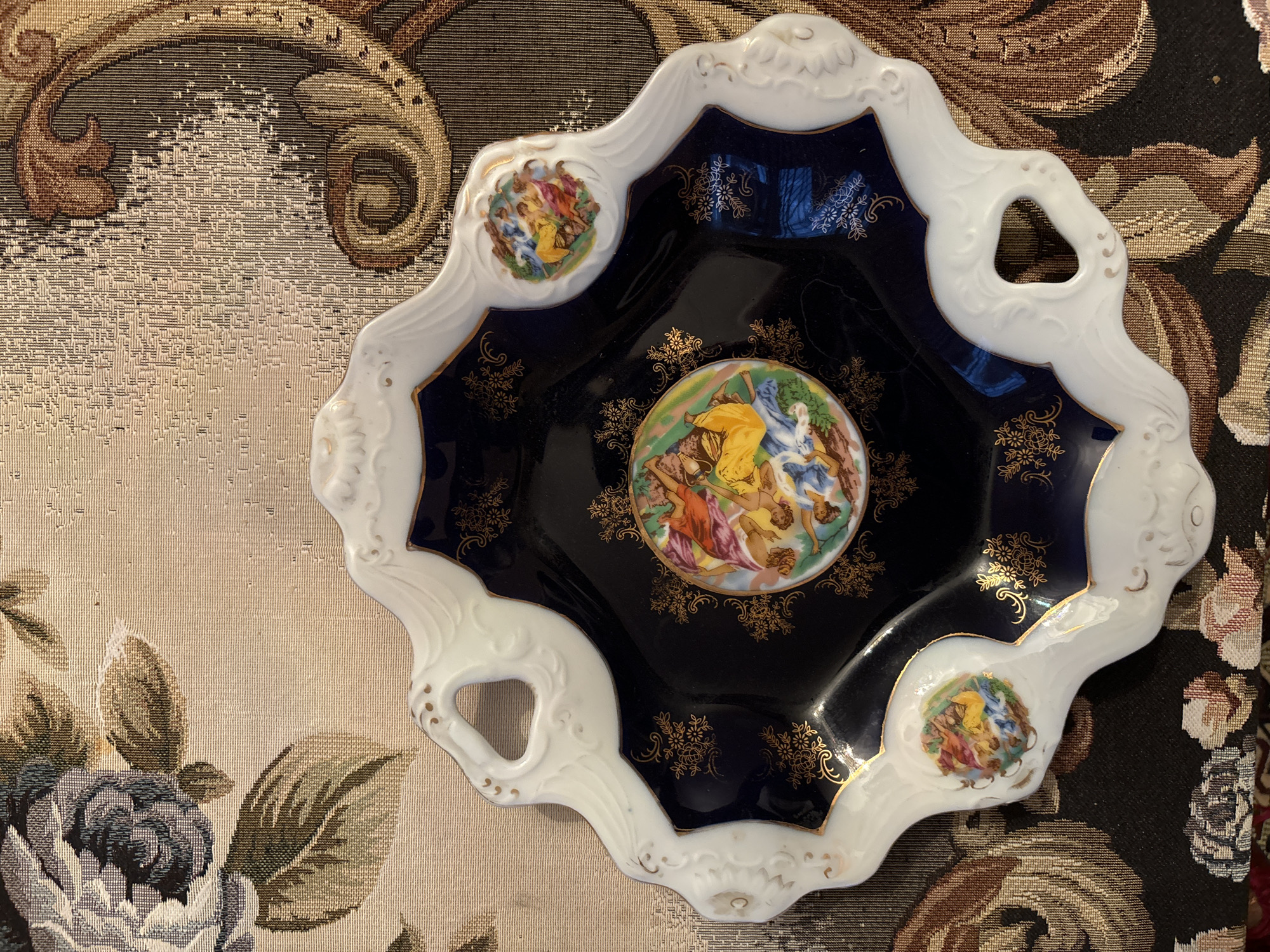 This is a top-down, medium shot of a decorative, dark blue and white porcelain plate with ornate gold trim. The plate has a scalloped edge and decorative cut-out handles. The center of the plate features a circular painting of several figures, possibly from classical mythology, in vibrant colors. The rim of the plate is decorated with smaller circular paintings of similar figures and gold floral accents. The plate is placed on a tapestry-like fabric that has a floral pattern in shades of beige, brown, and dark gray. The lighting is even, suggesting an indoor setting. No text is visible in the image.