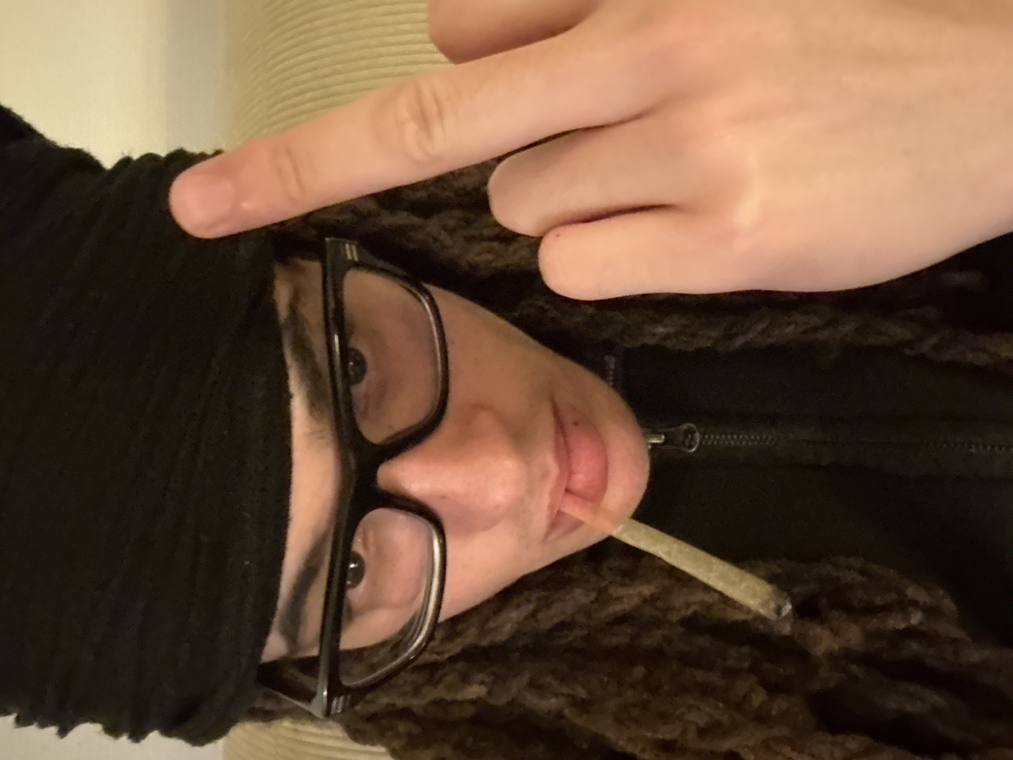 A person wearing a black head covering and large black-rimmed glasses is visible from the nose up, holding a lit cigarette in their mouth and with a hand raised over their head. They appear to be wearing a black zippered jacket and have brown dreadlocks. The background is a neutral color. There is no evidence to suggest the photo was taken in Paris, France.