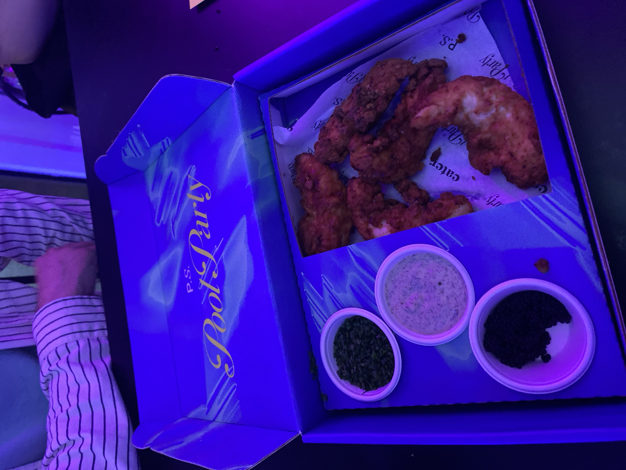A box of fried chicken tenders and three dipping sauces sits open on a dark surface. The box is blue with the words "Pool Party" in yellow script. The chicken tenders are golden brown and appear to be resting on a sheet of paper with the words "Party Cater" printed on it. The three dipping sauces are in small white cups. The sauces appear to be green, white, and black. A person wearing a striped shirt is partially visible on the left side of the image.
The location is not discernible from the image.