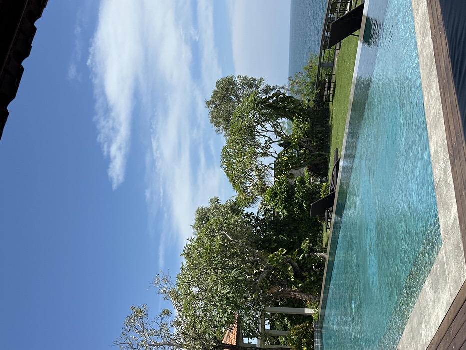 The image depicts a serene outdoor scene, likely a resort area, located in Uluwatu, Indonesia. In the foreground, a dark brown roof overhang partially frames the top right of the composition. Below this, the edge of a rectangular infinity pool with turquoise water is visible, featuring a light grey stone coping and a dark wooden deck surround on the left and bottom.
Beyond the pool, a well-maintained green lawn extends towards the horizon. Two dark-colored sun loungers are positioned on this lawn; one is located centrally behind the pool, and another is partially visible on the far right. A dense line of tropical trees and shrubs with vibrant green foliage forms a natural barrier behind the lawn. To the far left, a structure with terracotta-tiled roofs and light-colored columns is partially obscured by trees.
The vegetation gives way to a clear view of the open ocean, characterized by calm blue waters extending to the horizon. The sky above is bright blue with scattered white, wispy clouds, indicating clear weather. No individuals are present in the scene, and no overt activities are depicted. The overall impression is one of tranquility and natural beauty.