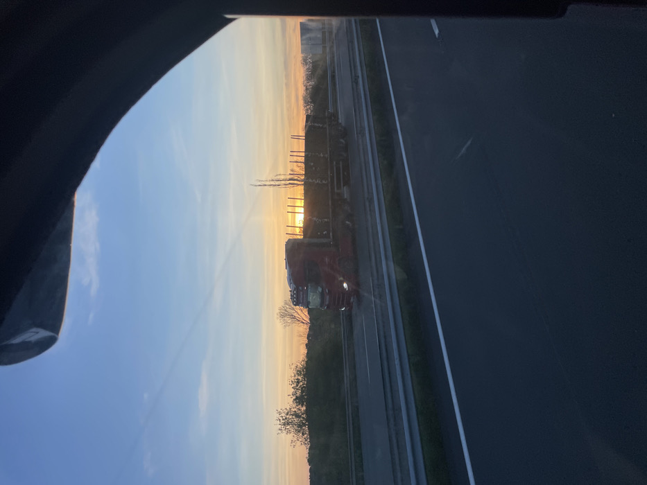 A red Scania semi-truck with a flatbed trailer, loaded with timber, is captured traveling on a multi-lane highway during sunset near Portel-des-Corbières, France. The truck moves from left to right, its headlights illuminated. The sun, a bright orange-yellow sphere, is visible at the horizon directly behind the truck, casting an orange hue across the lower sky that fades into light blue overhead. The highway features distinct white lane markings and a metal guardrail separating the lanes. Sparse trees and vegetation are visible along the distant horizon. The image is framed by a dark, irregular object in the upper and right sections, indicative of a vehicle's interior window frame from which the photograph was taken.