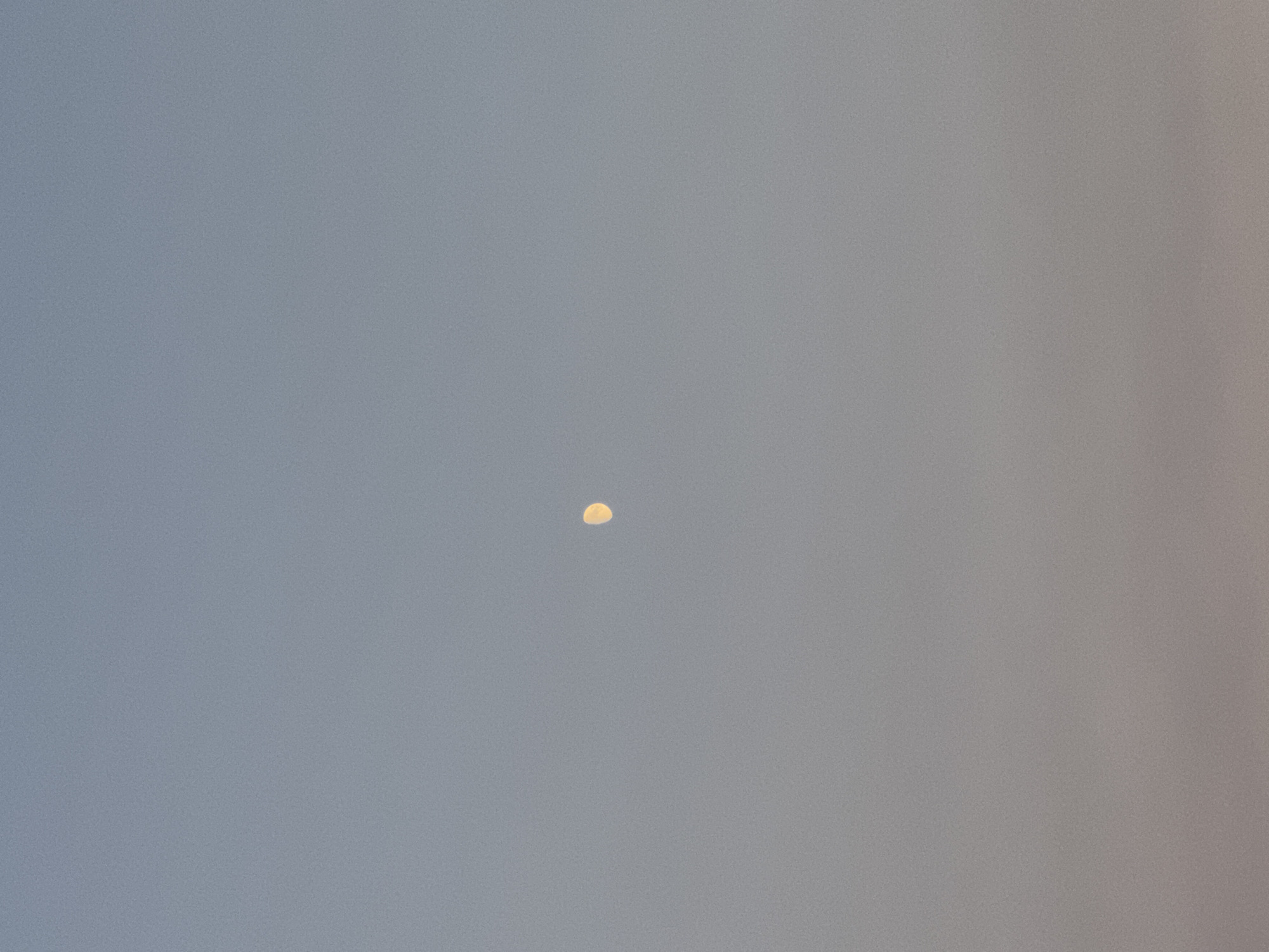The image depicts a clear sky with a gradient of blue and light purple hues, suggesting either early morning or late evening. In the upper middle portion of the frame, a small, crescent-shaped moon is visible, illuminated with a soft yellow light. The lower part of the image is darker, with hints of a faint, rosy glow near the horizon, possibly indicating sunset or sunrise. There are no discernible people, buildings, or specific landmarks visible, making it impossible to determine a precise location within Zagora, Montenegro, or any other city. The scene is serene and minimalistic, focusing solely on the celestial body against the backdrop of the sky.