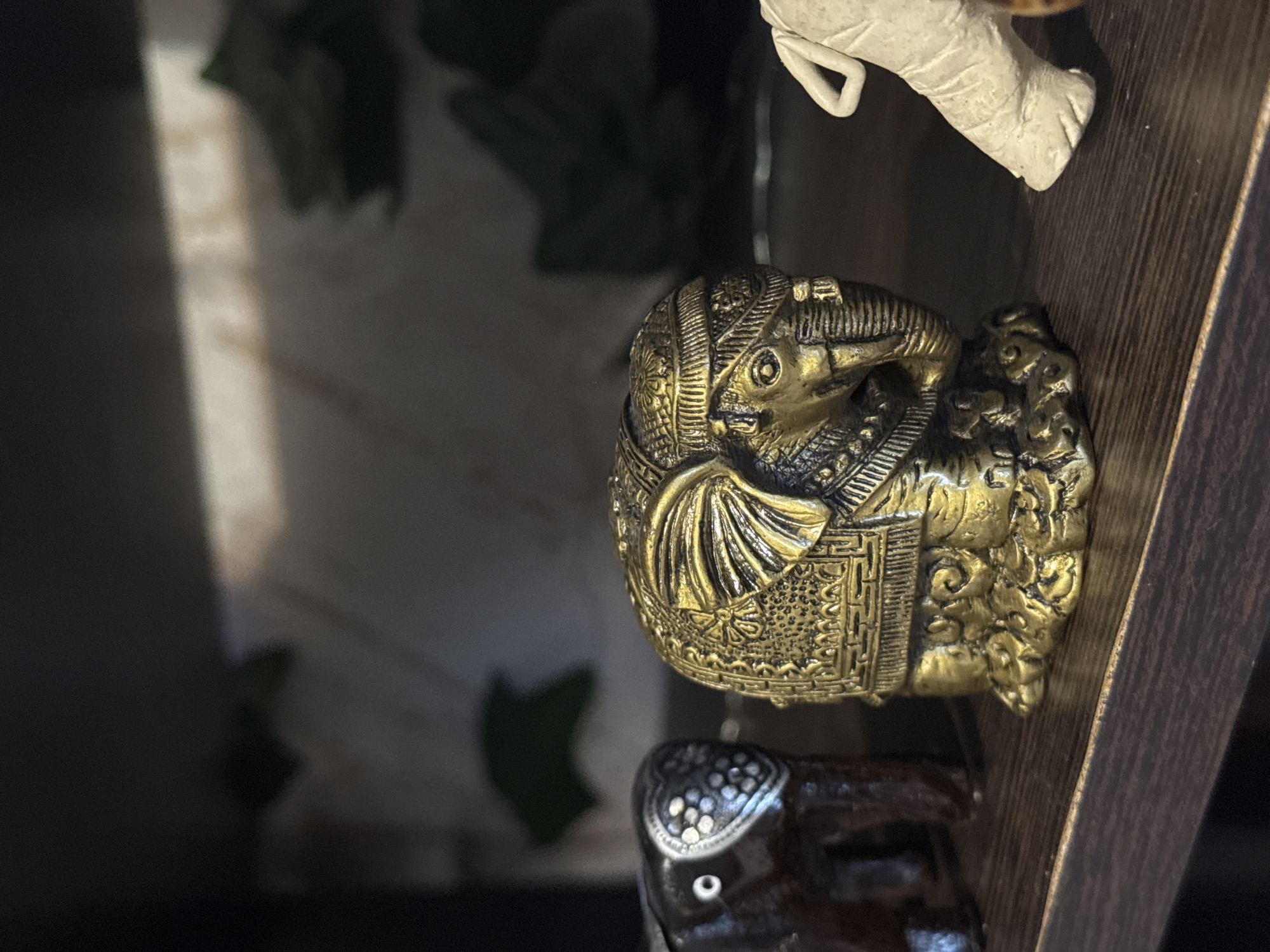 The image depicts a close-up of decorative objects displayed on a dark wooden shelf. A golden-colored, intricately detailed figurine of a bird, possibly an ibis or related species, is in focus. The figurine is adorned with patterns and designs. Above the bird, a portion of a white figurine, possibly an animal's leg, is visible. Below the bird, part of a dark, ornate object is also seen. The background is blurred, revealing hints of foliage and the edge of a surface.
The location in the city of Kladovitsy, Russia cannot be determined.