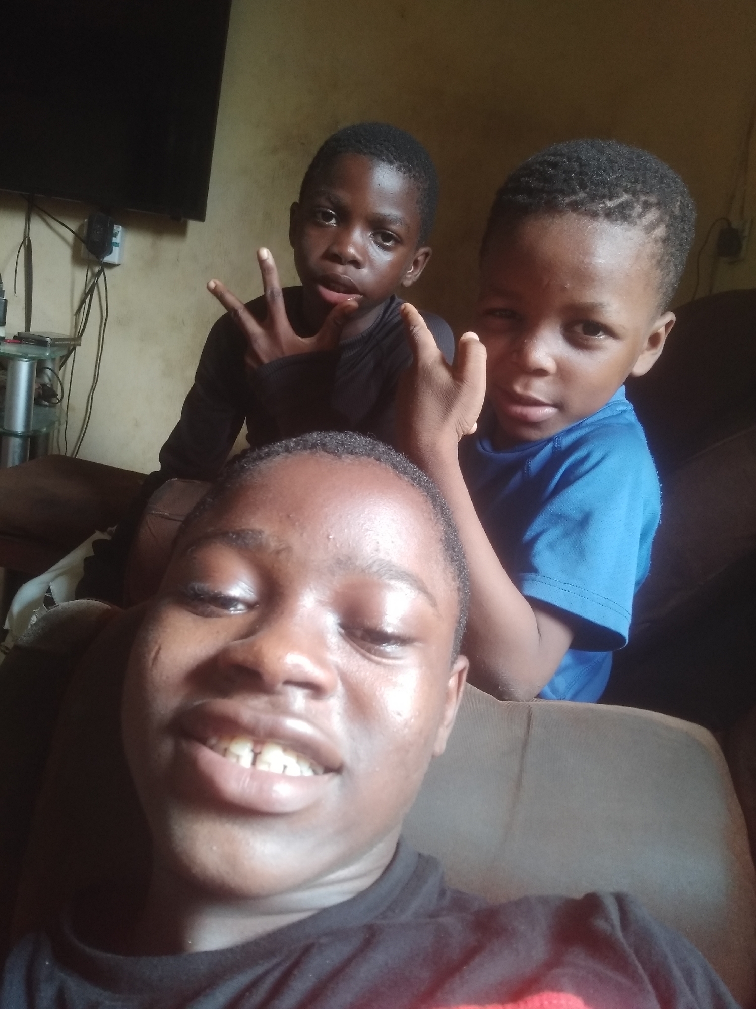 This image captures three young Black males posing together indoors, likely in a home in Shangisha, Nigeria.
In the foreground, a young male is positioned close to the camera, looking upwards with a broad smile that reveals his teeth. He is wearing a dark t-shirt and his face appears shiny, possibly from sweat or oil.
Behind him, to the left, another boy in a black long-sleeved shirt is looking directly at the camera, holding up three fingers in a gesture often associated with peace or a specific group. His expression is neutral.
To the right, partially obscured by the foreground subject, a third boy in a blue t-shirt is also looking towards the camera. He is making a similar hand gesture with his right hand, though it is less distinct. His hair is short and curly.
The subjects appear to be seated on a light brown, upholstered piece of furniture, likely a sofa. The background shows a light-colored wall. In the upper left corner, a large, dark flat-screen television is mounted on the wall. Below the TV, a small table or stand holds various electronic devices and a tangle of cables and wires, indicating a typical home environment.
The overall mood is casual and informal, suggesting a relaxed moment being captured. No discernible text is visible in the image.