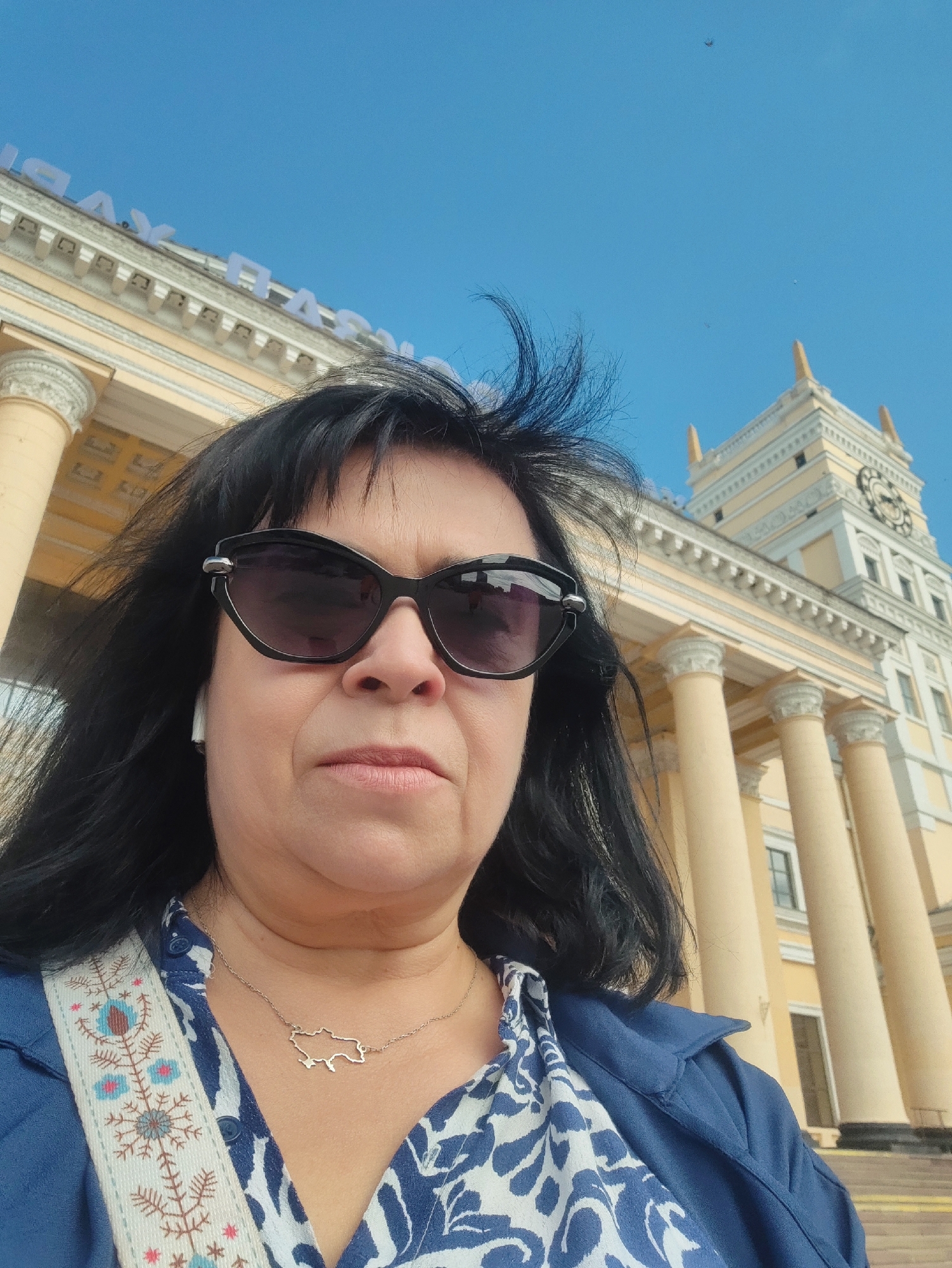 A woman with black hair and sunglasses is taking a selfie outdoors. She is wearing a blue jacket over a white and blue patterned shirt, and a necklace shaped like a map. Behind her is a large building with columns and a clock tower, and the sky is clear blue. The building has some text on its facade.