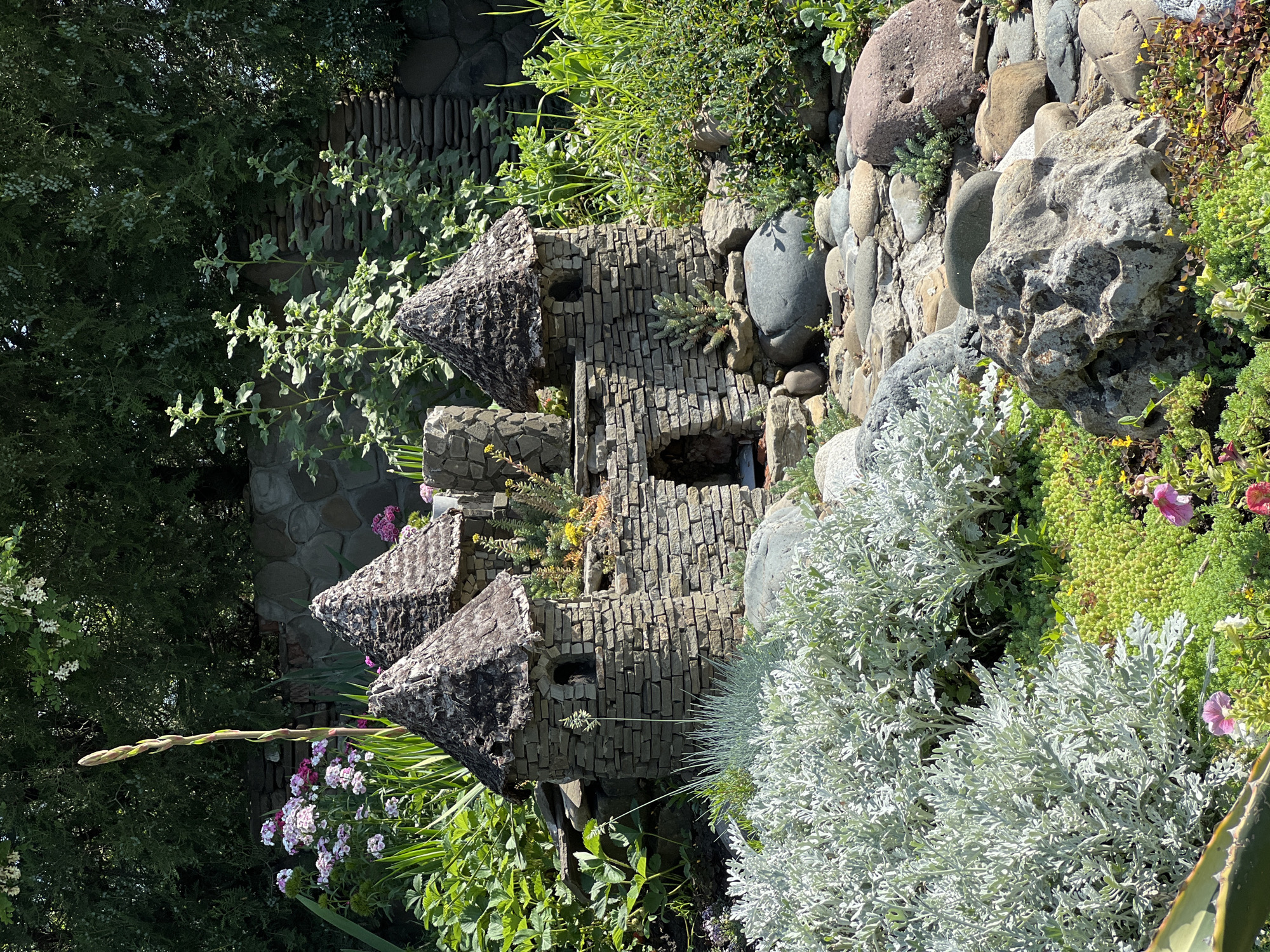 The image shows a miniature stone castle nestled among lush greenery and rock formations, suggesting a decorative garden feature. The castle is built with stacked, rectangular stones, featuring three distinct turrets with cone-shaped roofs also constructed from stone. Small openings resemble windows in the castle structure. The garden surrounding the castle is vibrant, with various plants including silvery-gray foliage, green shrubbery, and flowers. Larger rocks and stones form a backdrop and border to the garden feature. The background includes a stone wall and additional greenery. The location is in Krasnodar, Russia.