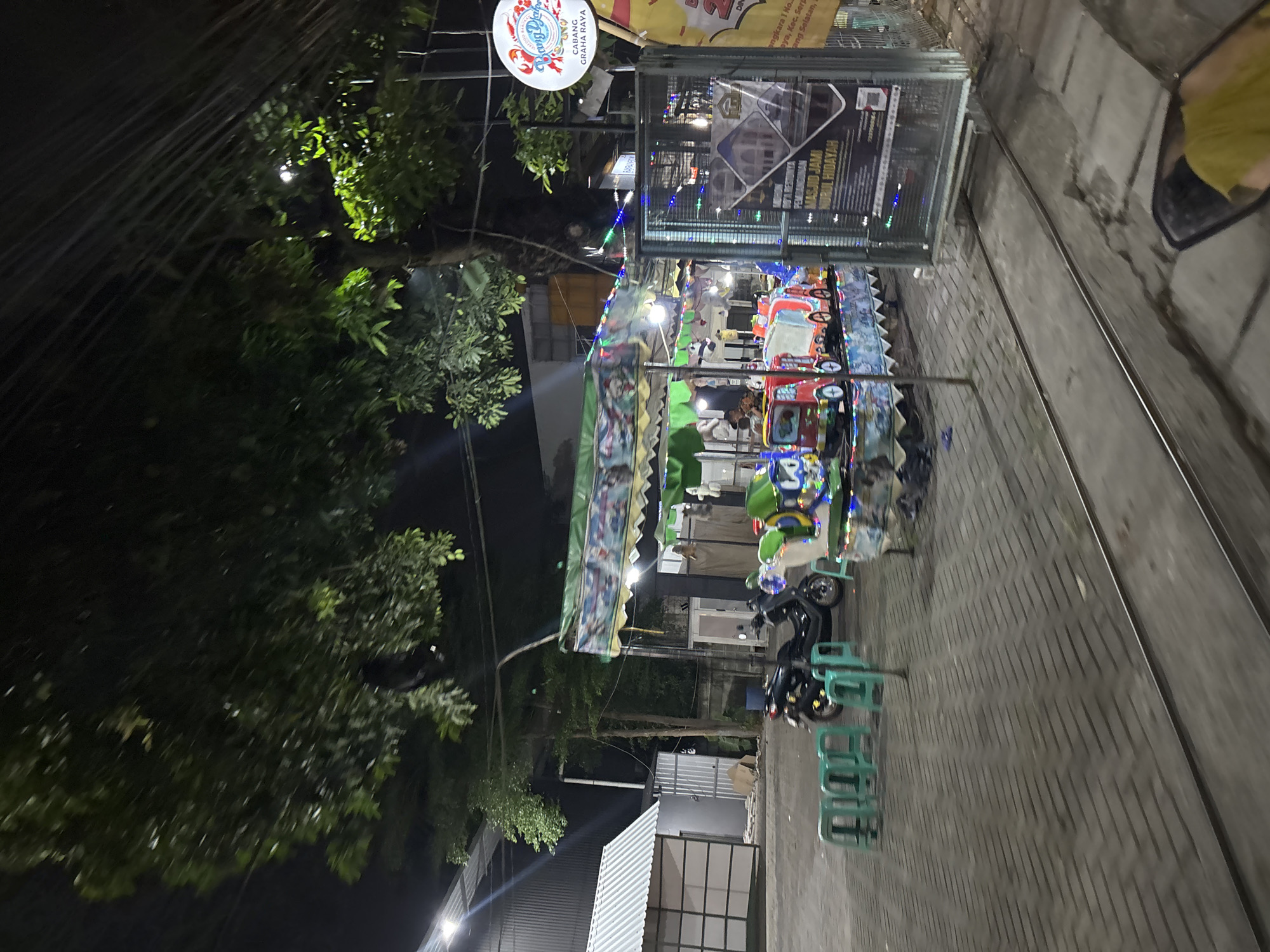 This image captures an outdoor scene at night, likely in South Tangerang, Indonesia. The primary focus is a small amusement ride area, possibly for children, with brightly colored toy vehicles. String lights are draped around the attraction, adding to the festive nighttime atmosphere. In the background, there are trees and some architectural elements, including a sign that appears to be for "Masjid Jami Nurul Hidayah" and mentions "Cabang Graha Raya," indicating a mosque branch. The ground is paved with patterned bricks, and there are metal tracks running along it, suggesting the amusement ride might follow these tracks. A black scooter is parked nearby, and several green plastic chairs are stacked, ready for visitors. The overall mood is casual and inviting, set against the backdrop of a clear night.