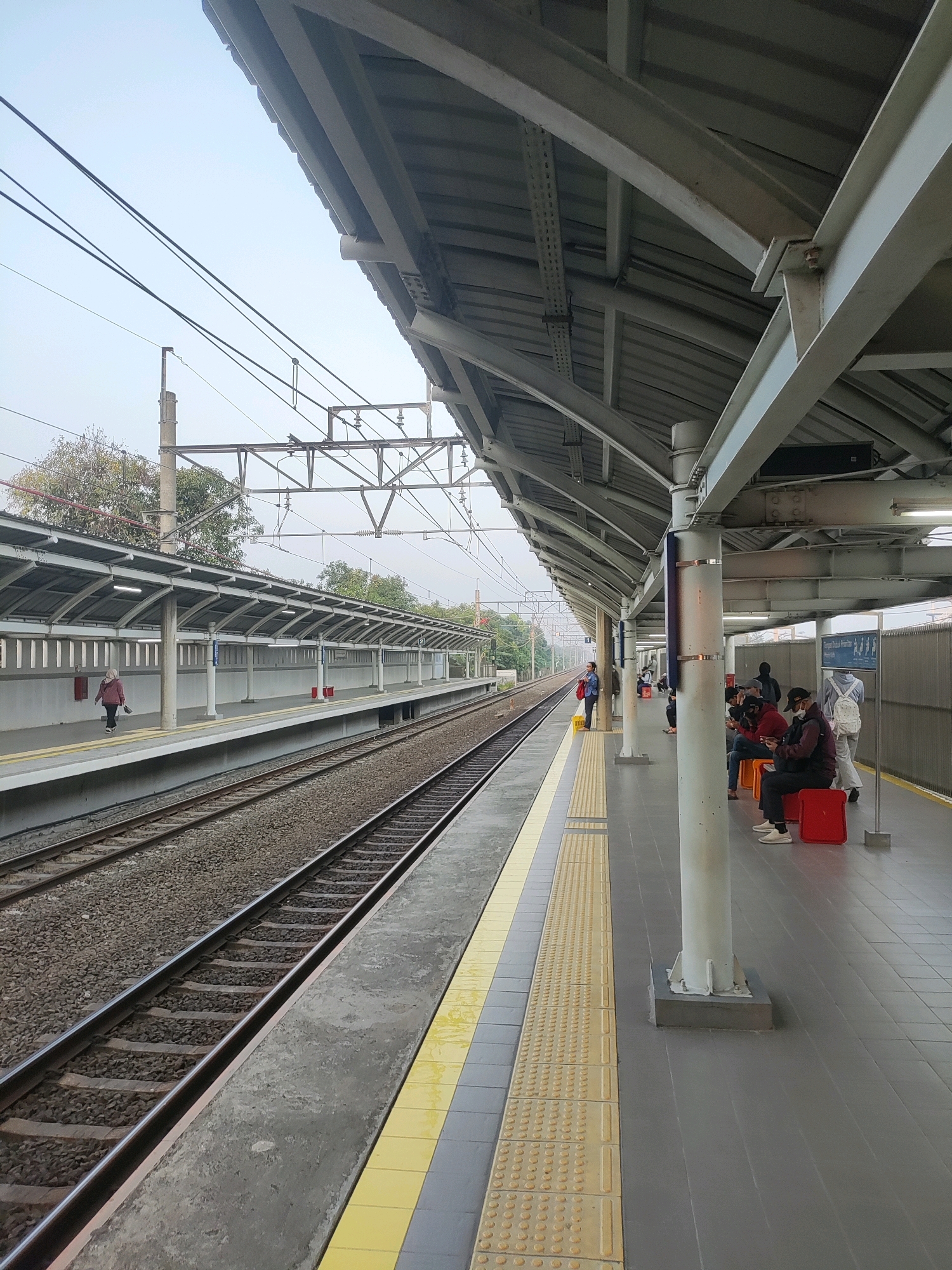The image depicts a train station platform in Telagamurni, Indonesia. A covered platform with a corrugated metal roof supported by white columns runs along the right side of the frame. Several people are seated on orange and red benches on the platform, some wearing face masks. A woman wearing a hijab and a maroon jacket walks along the platform on the left side, away from the camera. Train tracks are visible to the left of the platform, with ballast and sleepers. Overhead power lines for the trains are strung across the sky. The platform is paved with grey tiles and features a yellow tactile paving strip along the edge. The environment appears to be overcast or foggy.