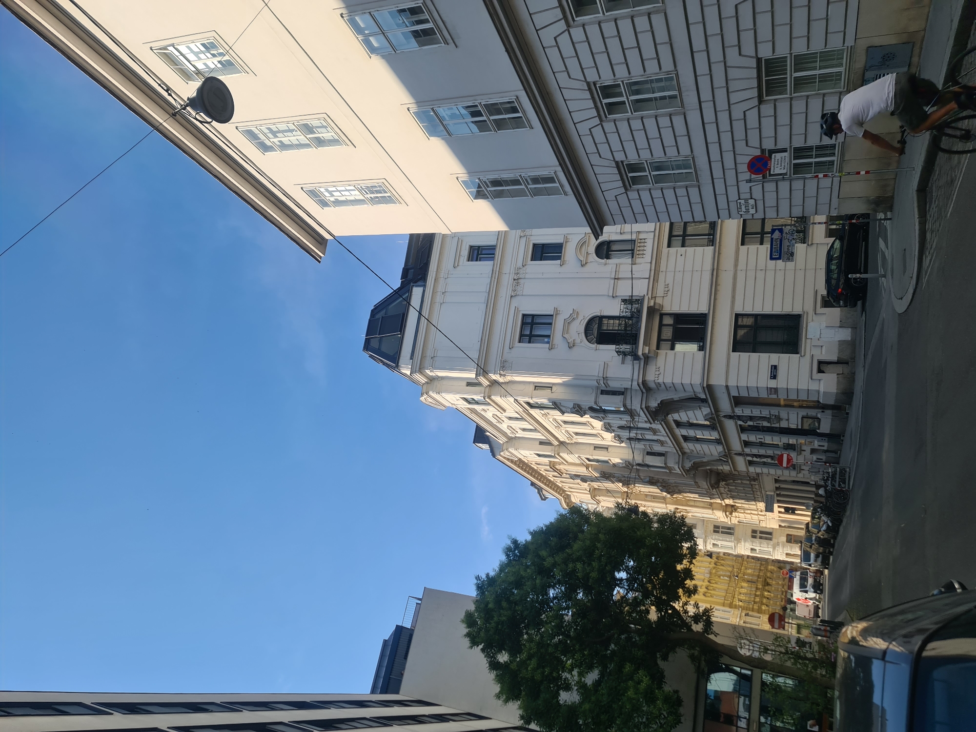 The image depicts a street scene in Vienna, Austria, under a clear blue sky. The viewpoint is from street level, looking up at the facades of several buildings lining a street. The architecture is predominantly classical, with ornate details on the upper floors and numerous windows.
On the right side of the frame, a light-colored building features a textured lower section and multiple large, multi-paned windows on its upper floors. A dark street lamp hangs from a wire suspended across the street, attached to this building. The building on the left is modern, with a flat facade and dark-framed windows. Between these two prominent structures, other buildings with varying architectural styles and colors can be seen, including one with a bright yellow facade further down the street.
A large green tree with dense foliage is situated to the left, partially obscuring the modern building. The street itself is paved, and several cars are parked along the right side. In the middle ground, a white van and other vehicles are visible.
A person is riding a bicycle in the foreground on the right side of the street, wearing a white t-shirt and dark shorts, with a helmet on. They are moving away from the camera. There are also street signs, including a blue sign indicating "U-BAHN" (subway) and a circular red and white traffic sign. The overall impression is of a quiet urban environment during the daytime.