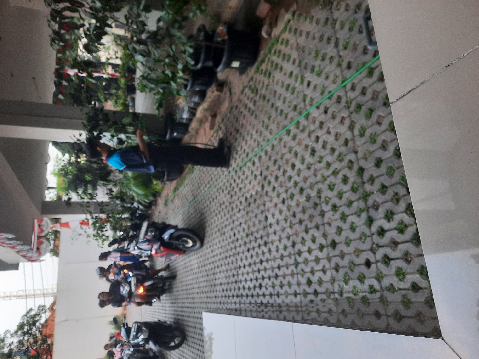 In North Jakarta, Indonesia, a person is seen watering plants with a green hose. The paved ground is made of interlocking concrete blocks with grass growing in the gaps. Several motorcycles are parked on the pavement. In the background, a building with red and white banners is visible, along with trees and potted plants. Some individuals are on or near motorcycles, suggesting a possible departure or arrival. The scene appears to be an outdoor area adjacent to a building, possibly a parking lot or a walkway.