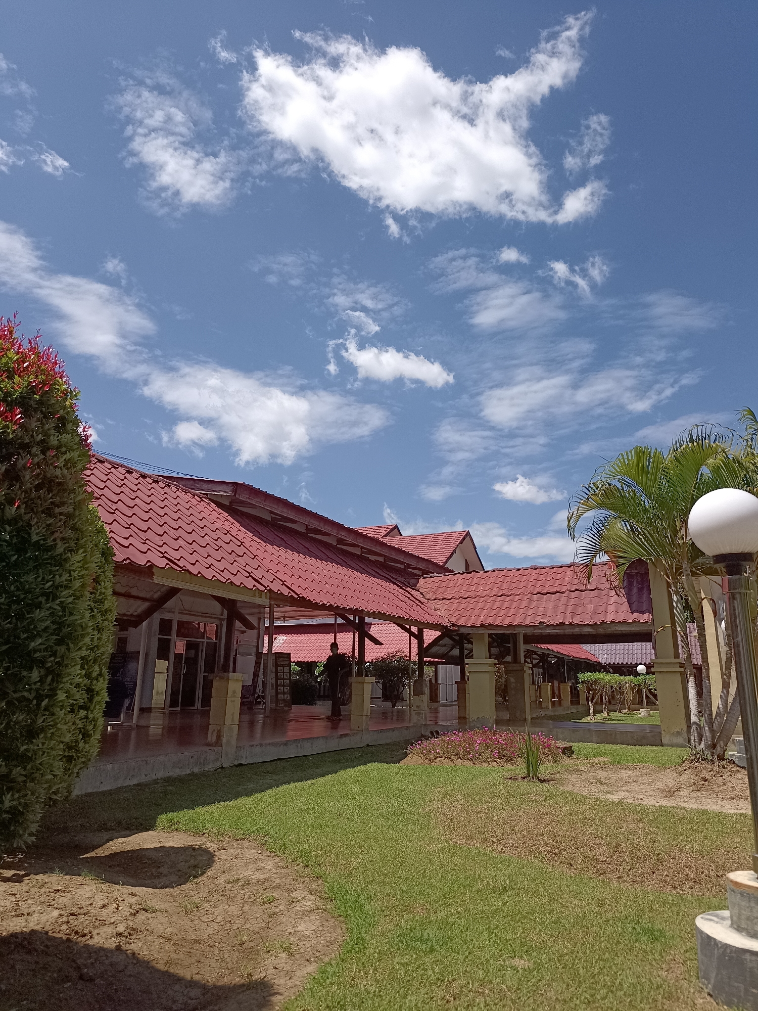 The image shows a building with a red tile roof under a blue sky with white clouds. In the foreground, there is a green lawn and a dirt path. To the left, there is a large green shrub. To the right, there is a lamppost and some palm trees. In the background, there are people walking under a covered walkway. The scene appears to be in a public area, possibly a park or a cultural center. The location is identified as Mesjid Reuntoh, Indonesia.