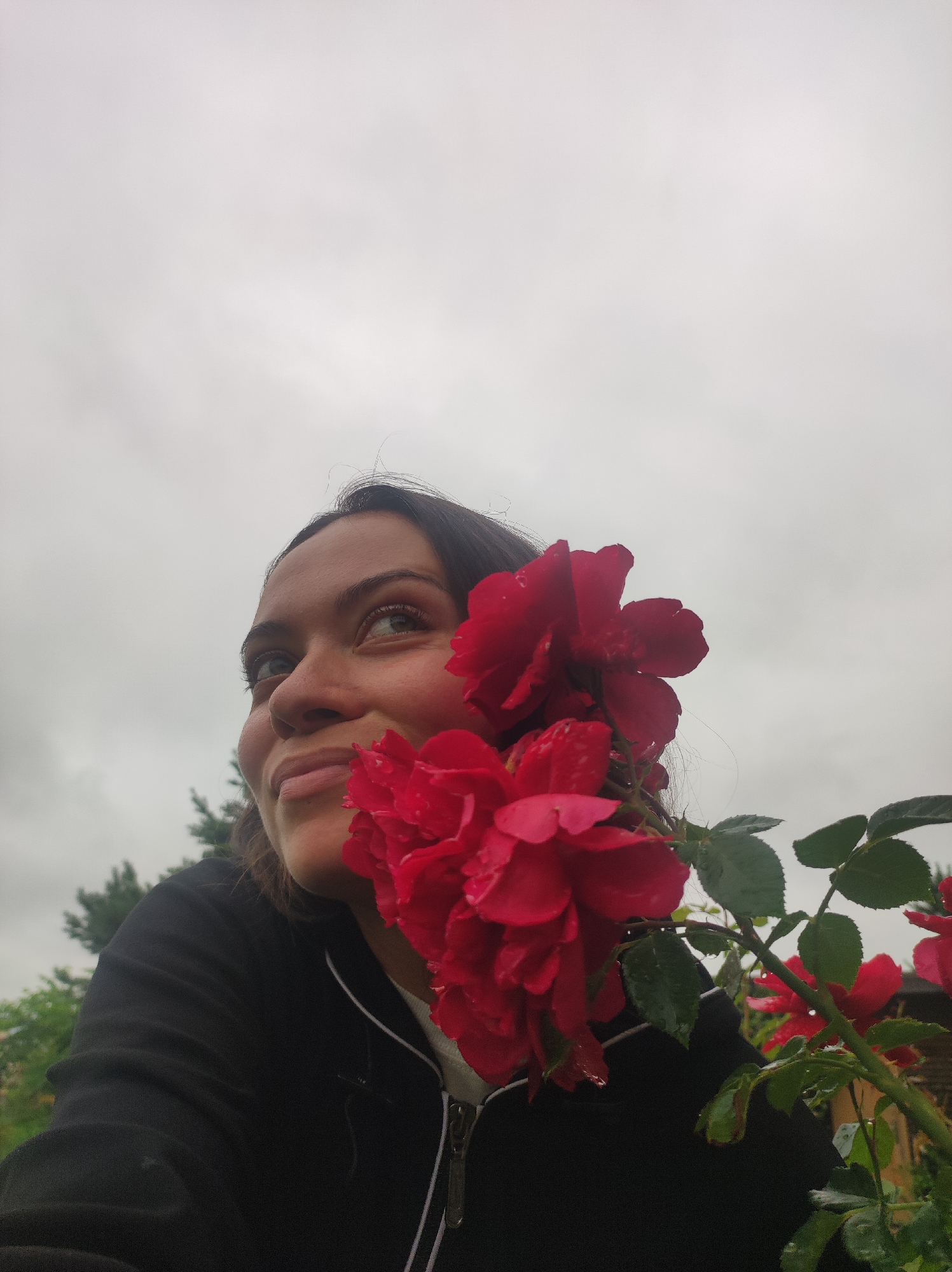 In this medium shot, a young woman with dark hair and brown eyes is looking upwards and to her left, with a slight smile. She is wearing a black zip-up jacket over a white shirt. In the foreground, a branch of red roses with green leaves is positioned over her right shoulder, partially obscuring her face. The roses appear wet, suggesting recent rain. The background is a blurred mix of greenery and a pale, overcast sky, indicating an outdoor setting, likely in a garden or park, under cloudy weather conditions. The overall mood is serene and contemplative. No visible text is present in the image.