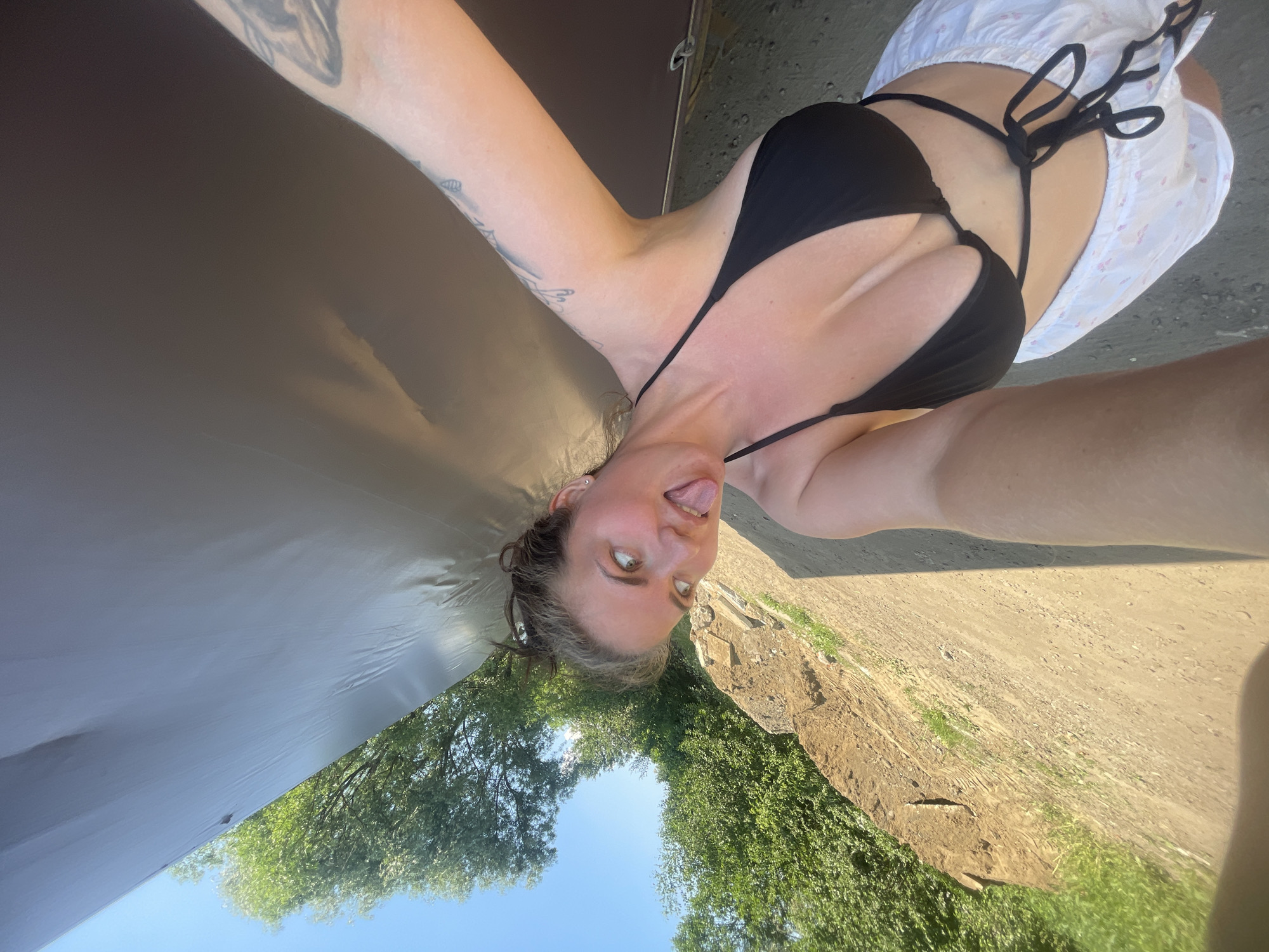 A young woman is taking a selfie outdoors. She is wearing a black bikini top and white shorts. Her hair is pulled back in a bun, and she is sticking her tongue out at the camera. The background shows trees, a dirt path, and a bright blue sky. The location is identified as Мосметаллоконструкция Онт, Russia.