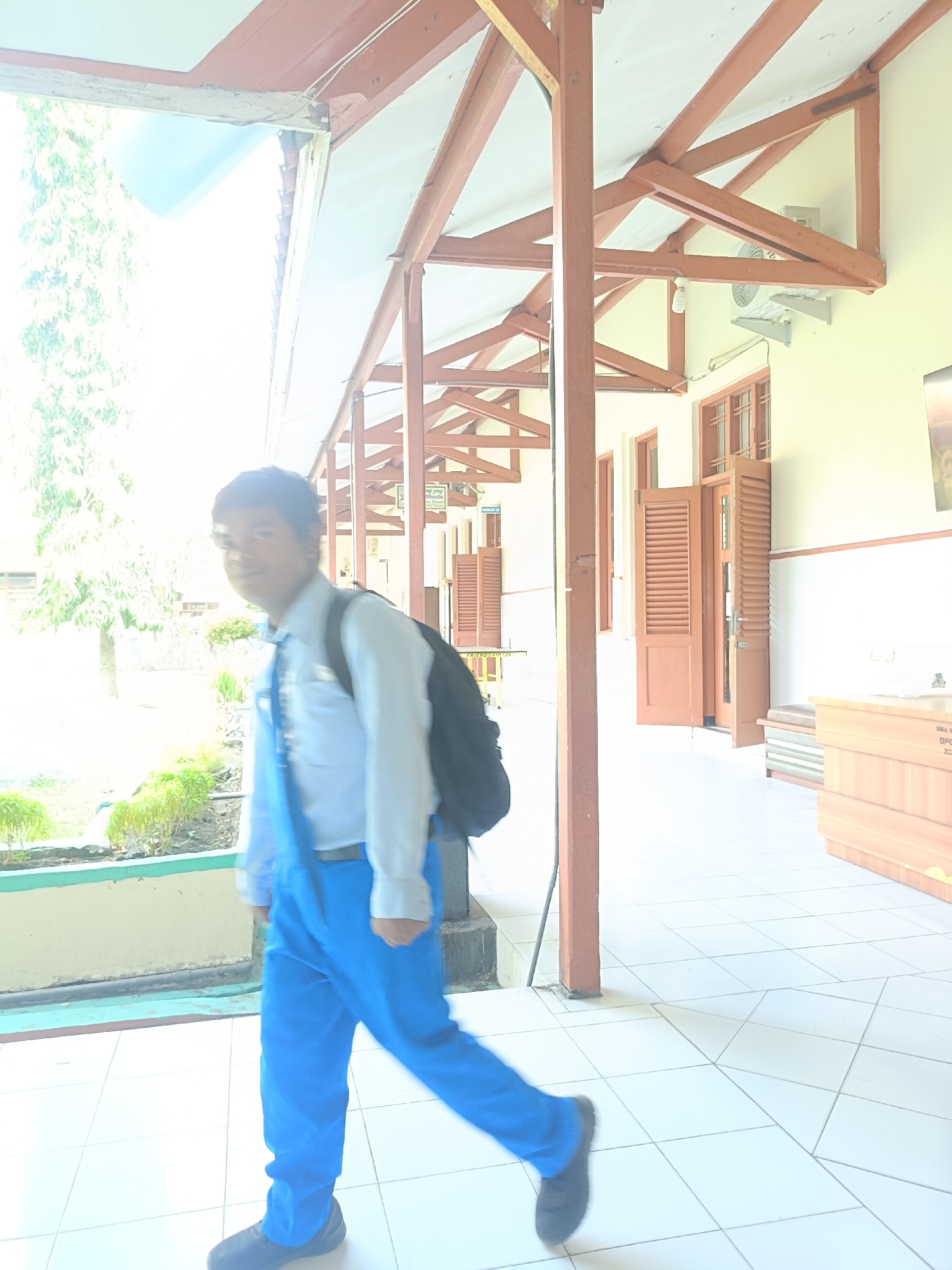 A young Indonesian boy wearing a light blue long-sleeved shirt, a dark blue tie, and bright blue pants walks on a tiled walkway in front of a building with a covered walkway. He is carrying a black backpack. The building has wooden beams supporting the roof and wooden doors with louvers. Green plants are visible to the left of the walkway. The environment appears to be outdoors, possibly in front of a school or other public building. The overall lighting suggests a bright day.