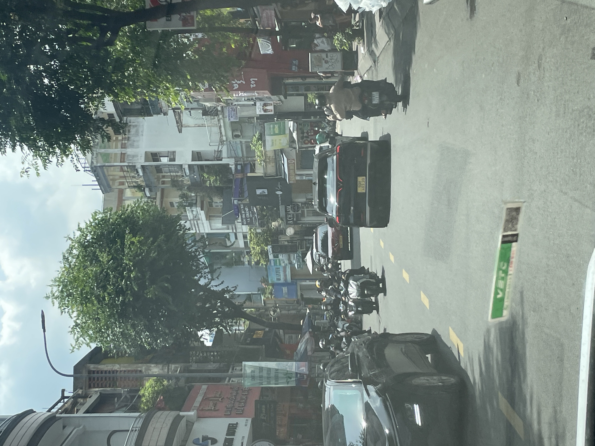 This image shows a street scene in Thủ Đức City, Vietnam. The street is lined with multi-story buildings, some of which appear to be shops or businesses. Trees are planted along the sidewalk, providing shade. Several motorcycles and a car are visible on the road. People are seen riding on the motorcycles. The sky is cloudy. The image appears to be taken from a vehicle moving through the street.